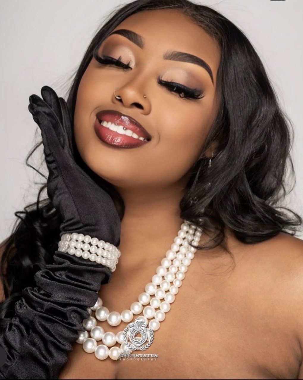 Woman with makeup, jewelry, and black gloves, smiling with eyes closed, touching face with gloved hand.
