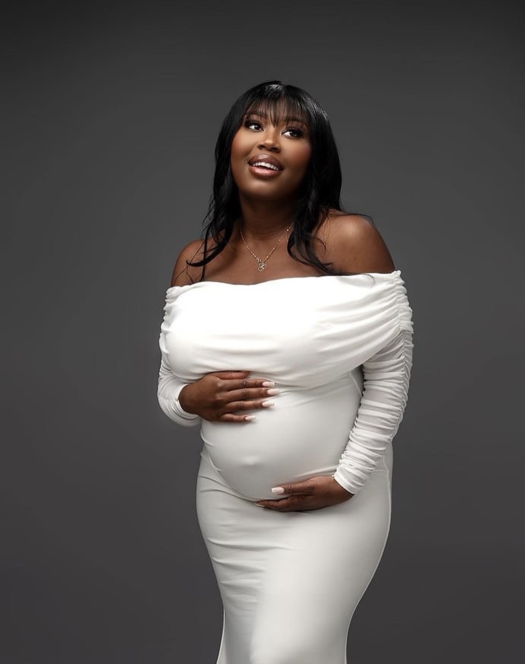 Woman in white off-shoulder dress with long sleeves, standing against a dark background, smiling, with one hand on her stomach.