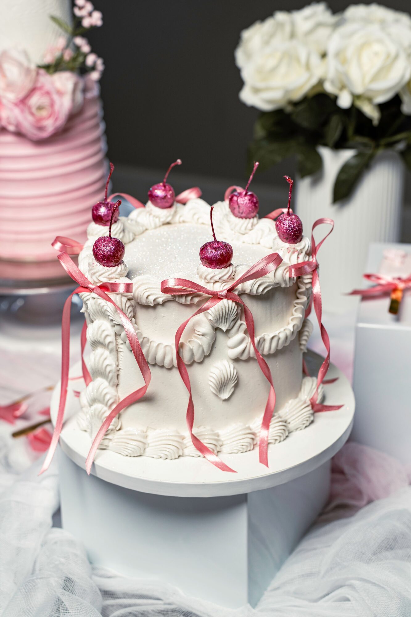 Decorative cake with cherries and pink ribbons, surrounded by flowers and tableware, on a white stand.