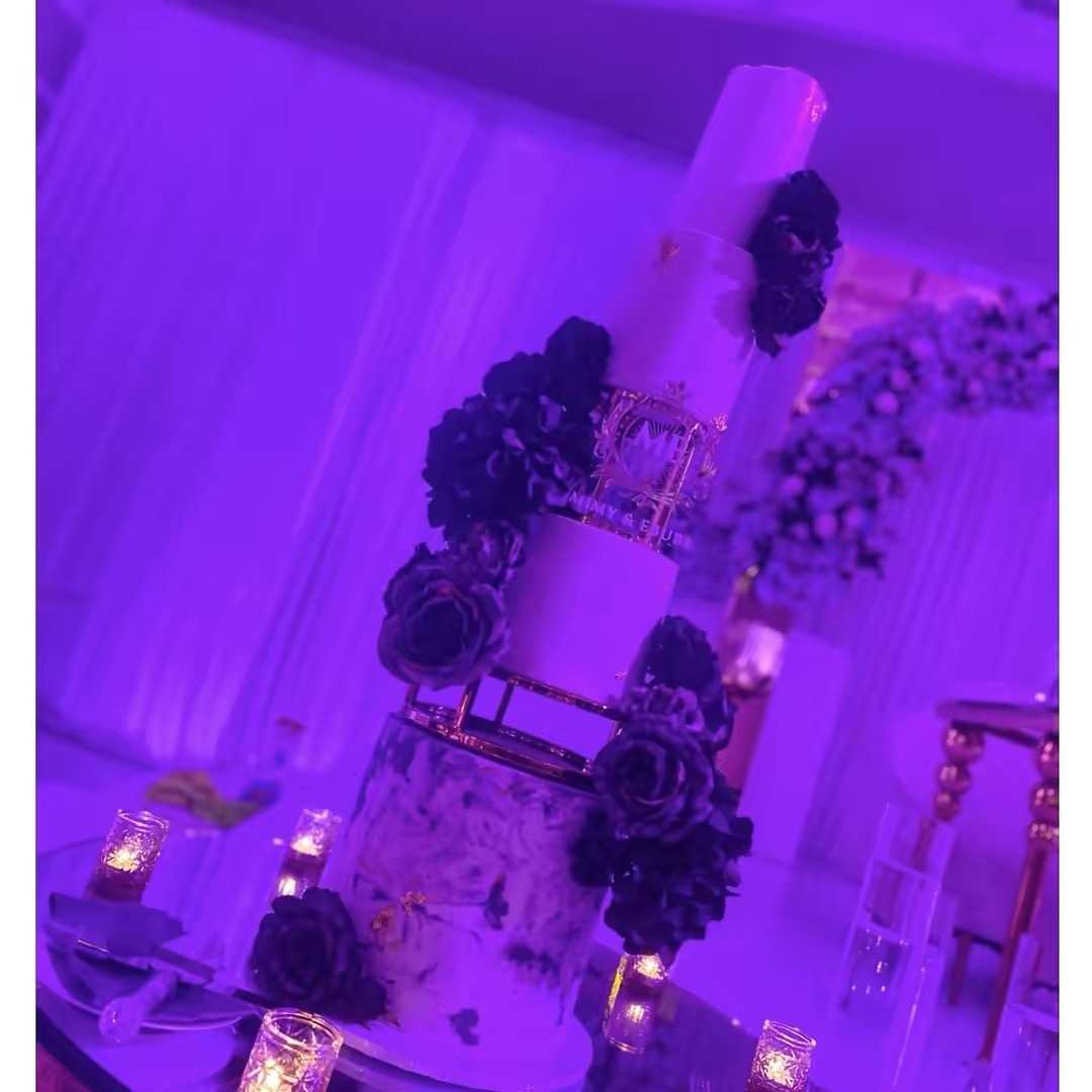 Upright wedding cake decorated with purple flowers and candles, set in an elegant venue with draped curtains and floral arrangements.