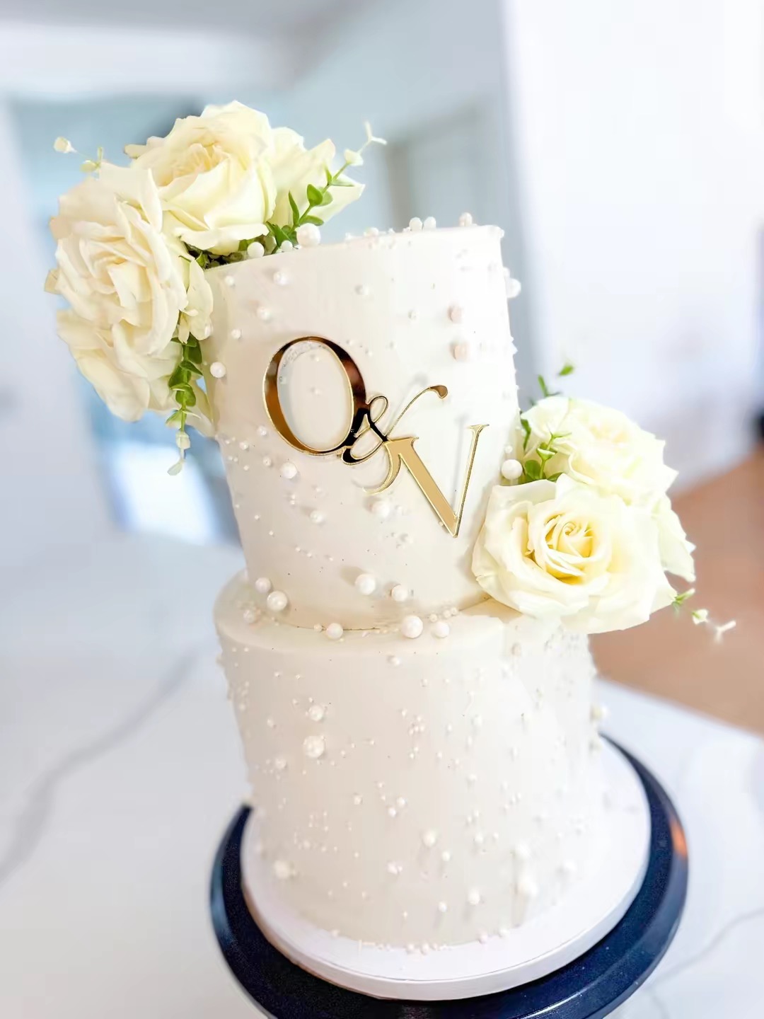 Two-tier white wedding cake decorated with white roses and small white pearls, with initials 'Q' and 'V' on the front.