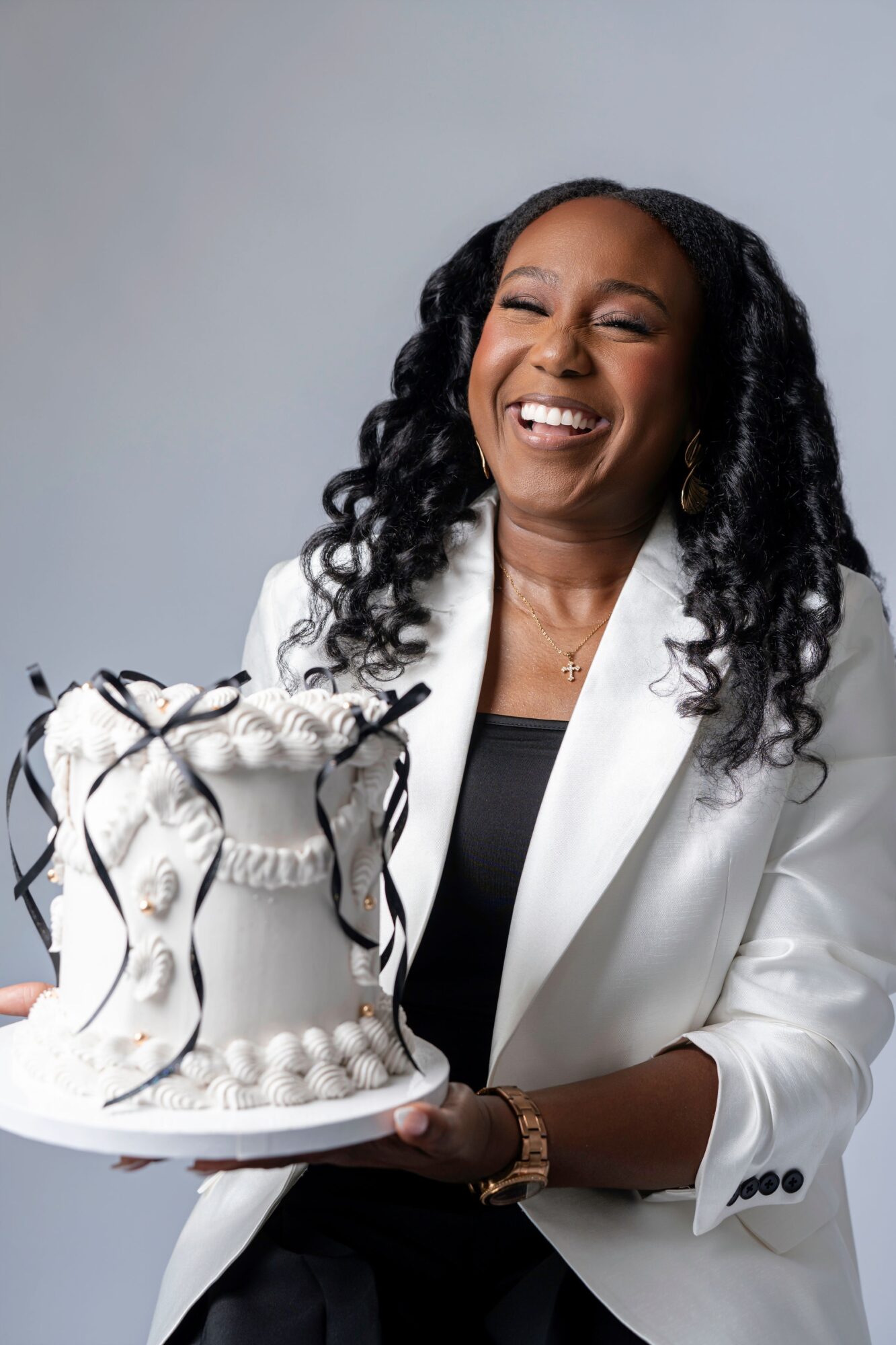 Woman with curly hair smiling, holding a decorated cake, wearing a white blazer and black top.
