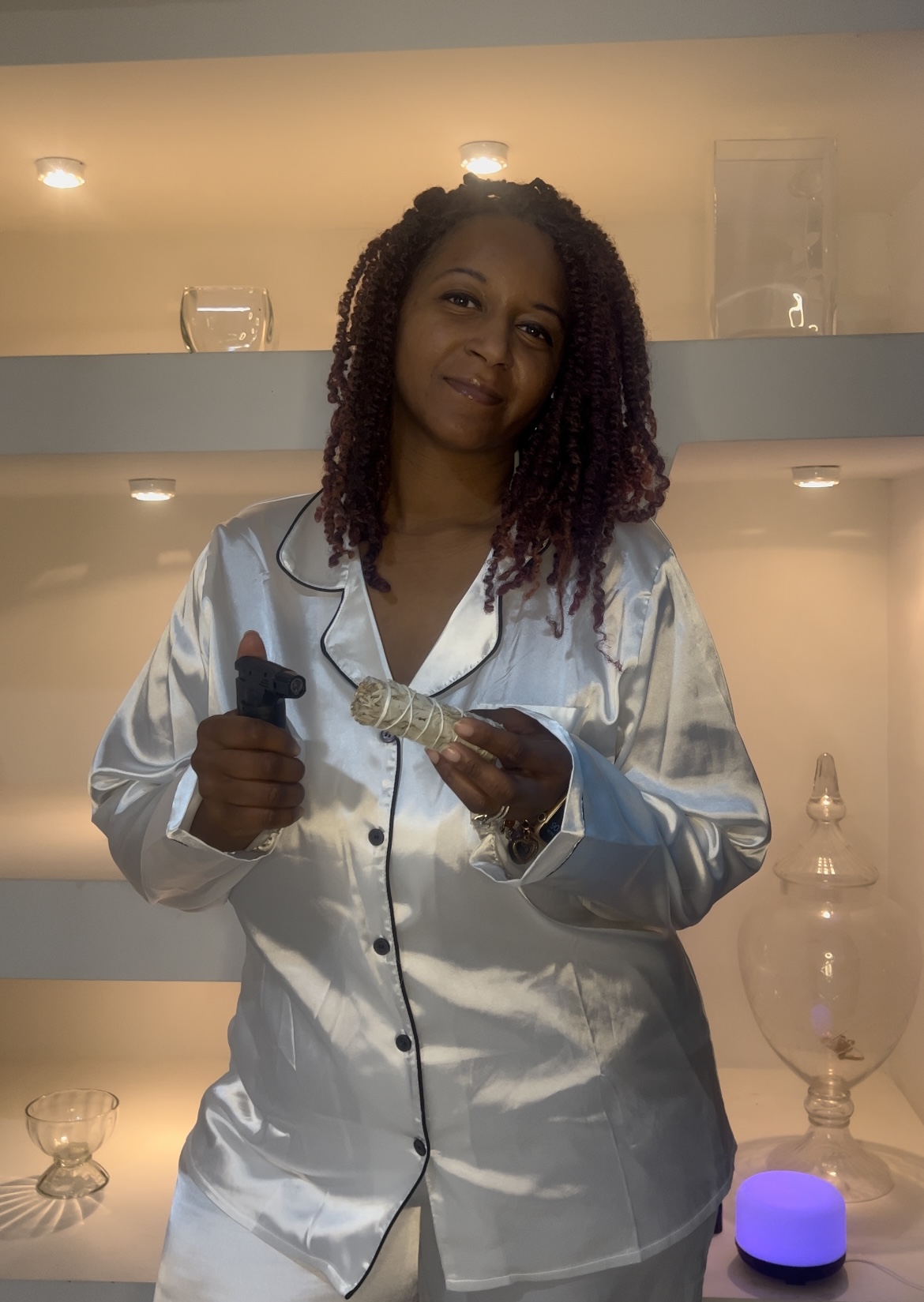 Woman in satin pajamas holding a microphone, smiling, standing in front of a shelf with decorative items, warm lighting.