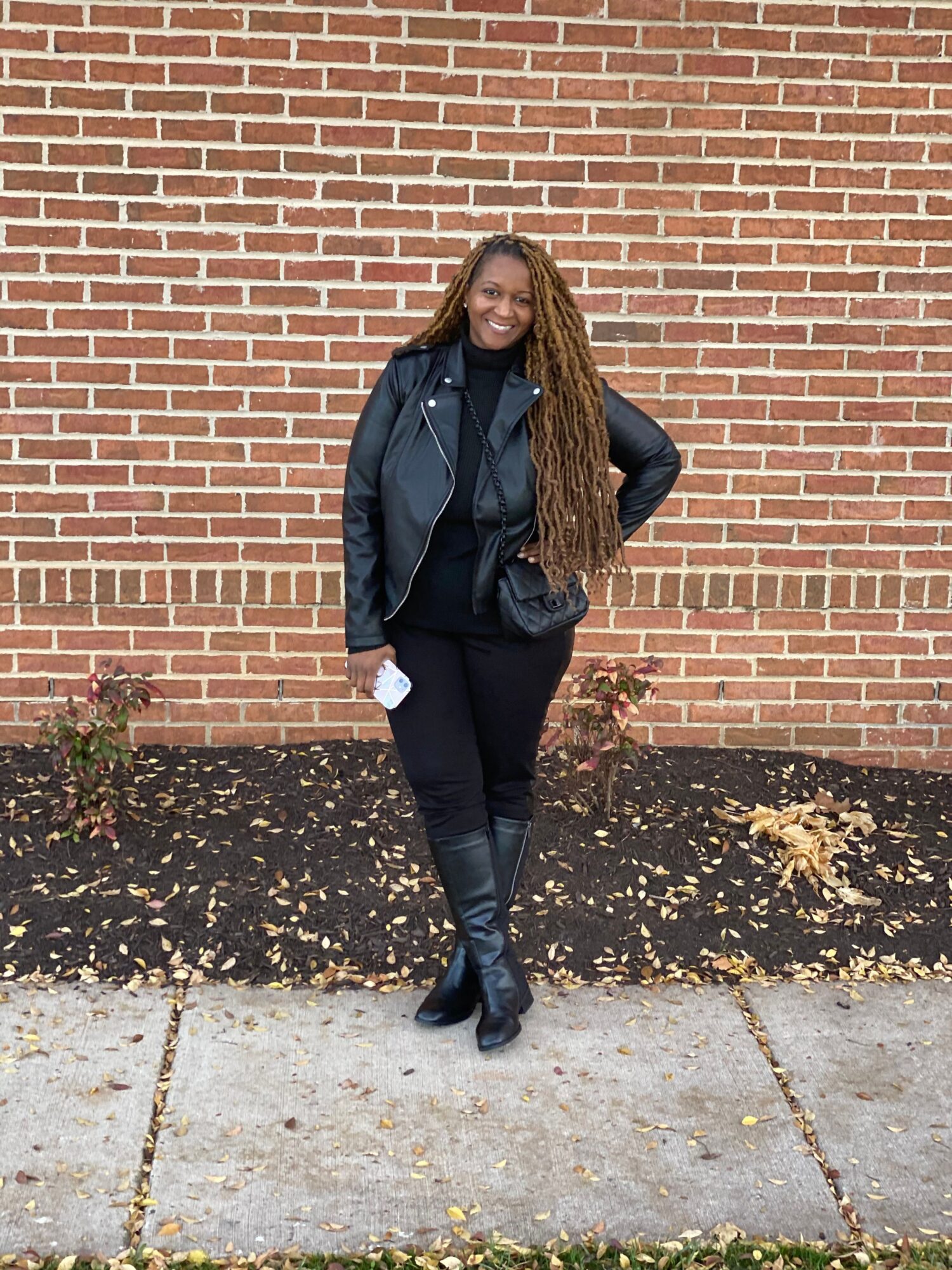 Woman with long hair, black jacket, black pants, and boots walking along a brick wall and sidewalk.
