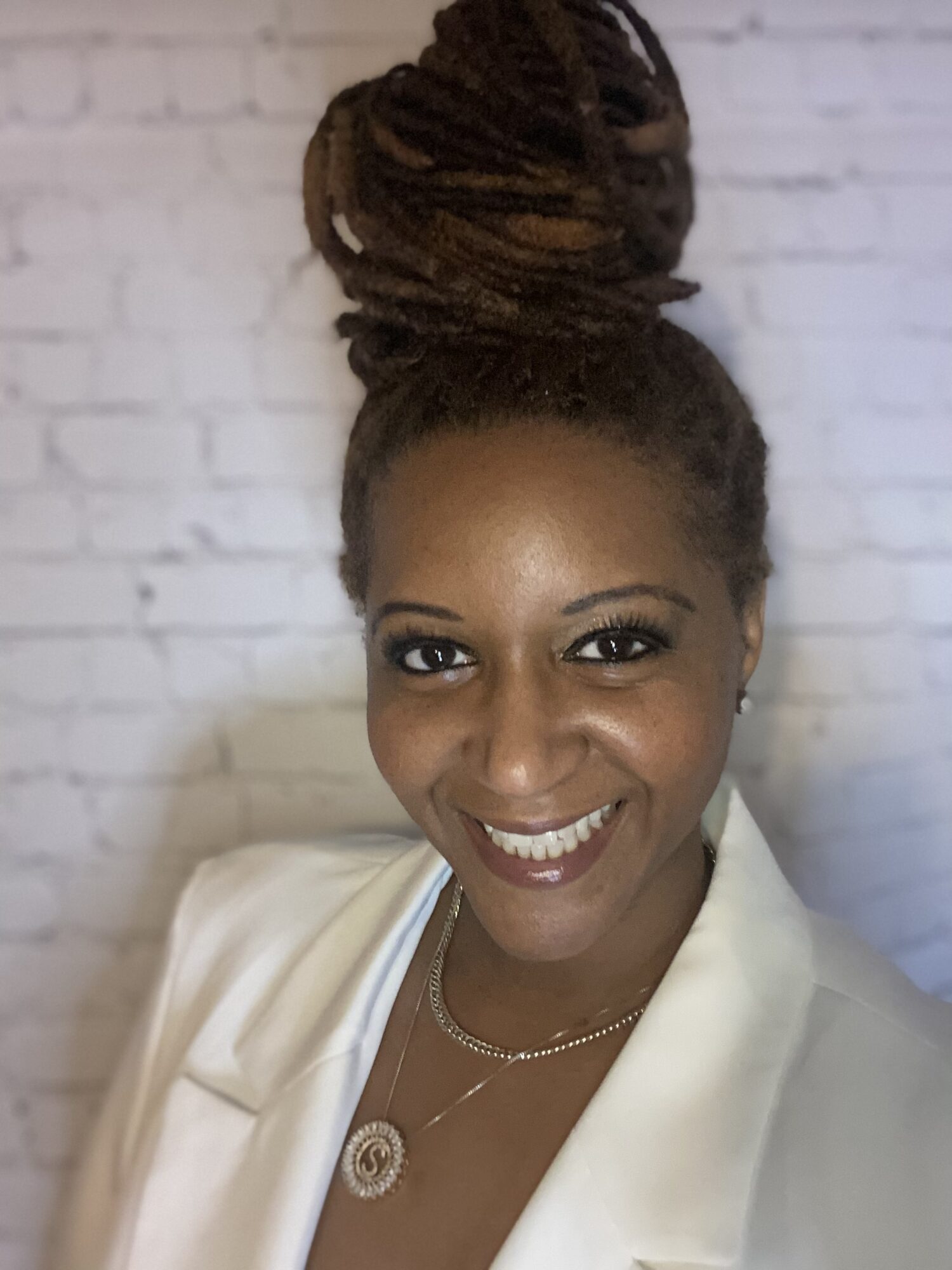 Portrait of a smiling woman with styled hair in a high bun, wearing a white blazer and a necklace, against a white brick wall.