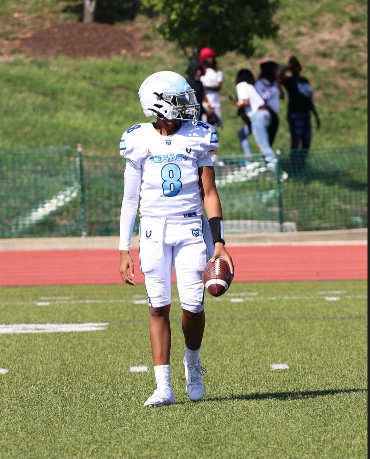 Football player in white uniform holding a football on a field with spectators in the background.