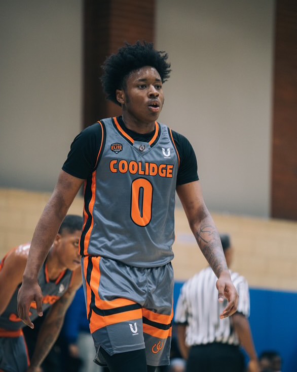 Basketball player in gray and black uniform with orange accents, number 0, standing on court. Two other players and a referee are in background.