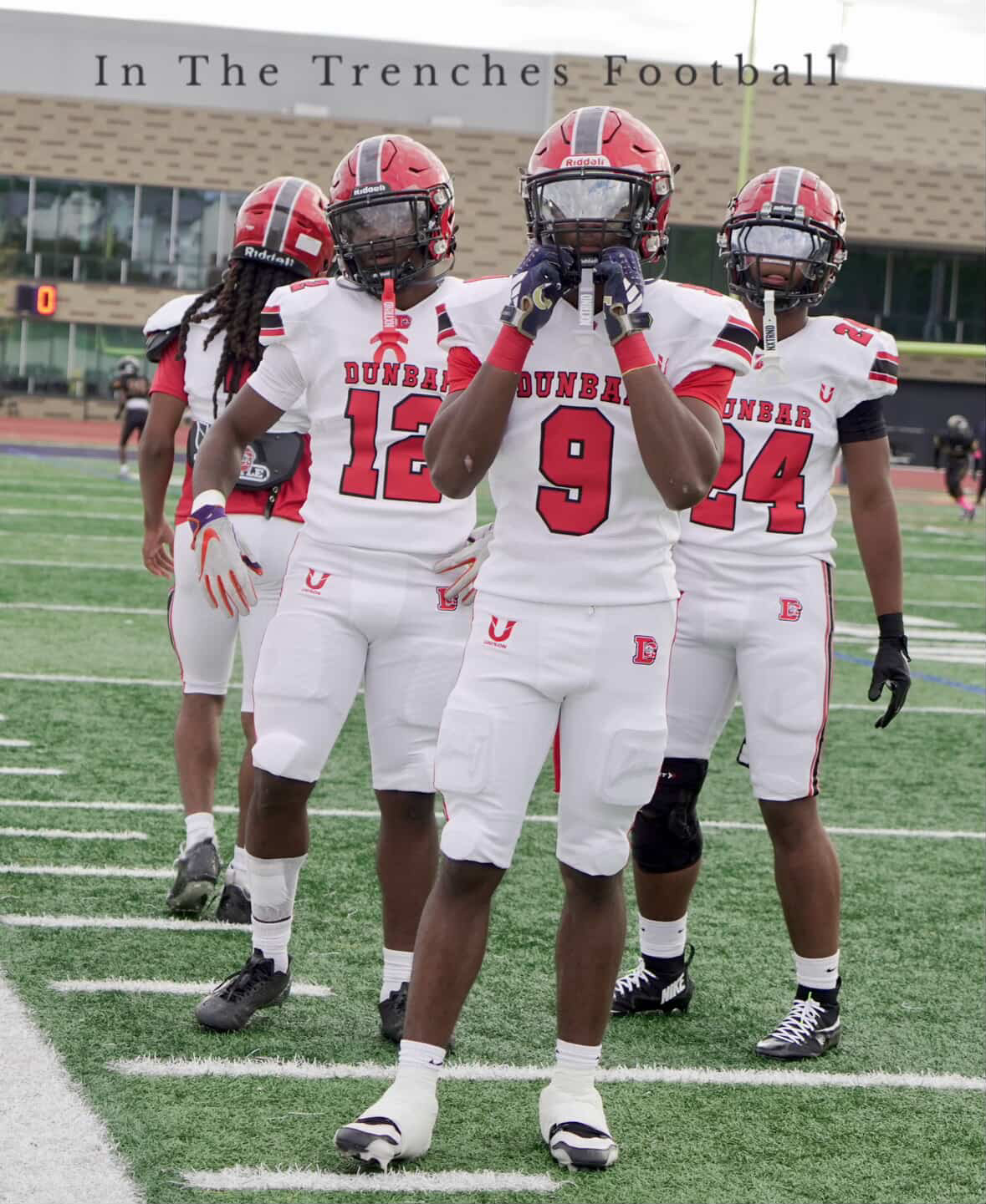 Four football players in white and red uniforms standing on a field, with a building in the background.