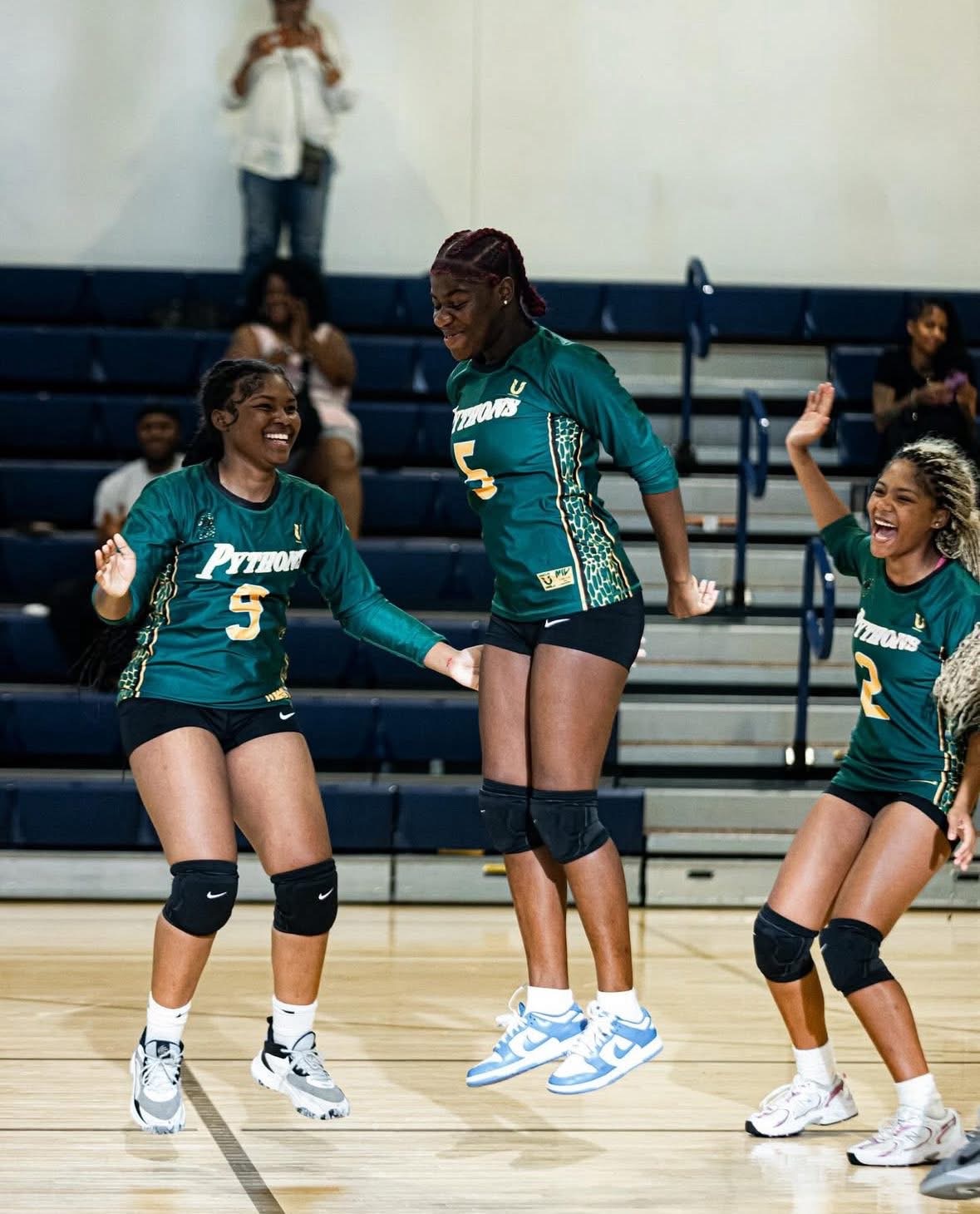 Three female volleyball players in teal uniforms celebrating on the court, with spectators in the background.