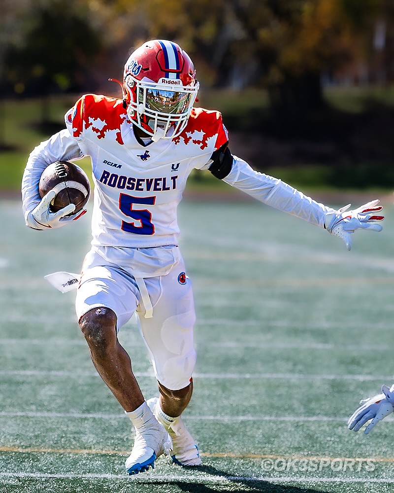 Football player in uniform running on field holding a football, with trees in background