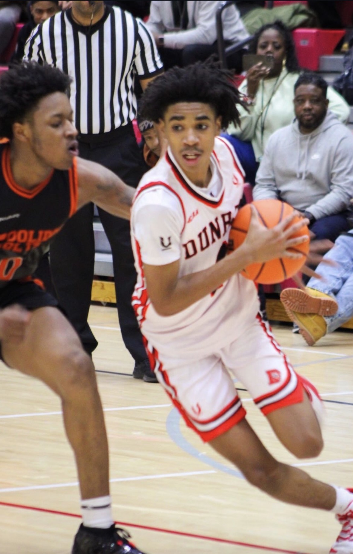 Basketball player in white jersey dribbling ball, opponent in black jersey reaching, spectators watching in background.