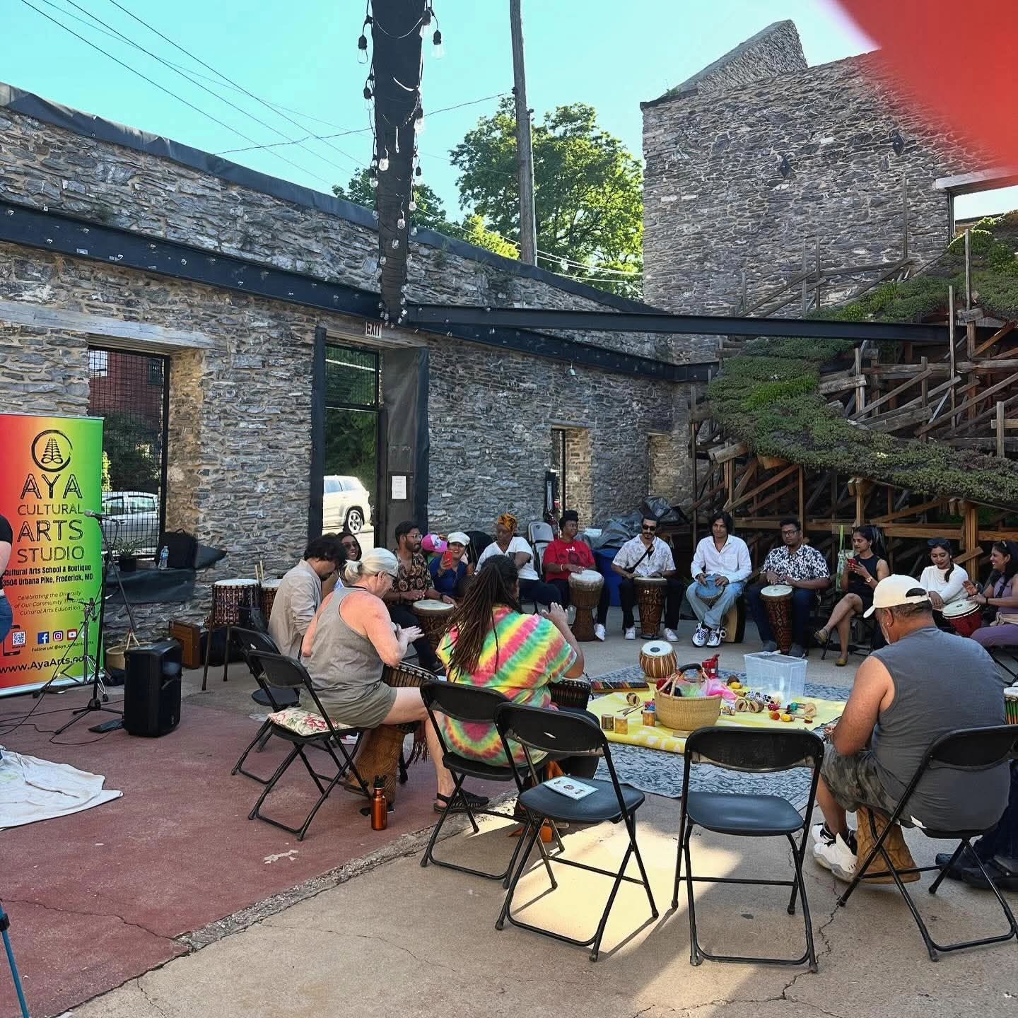 Group of people sitting outdoors in a courtyard with brick and stone buildings, some trees, and a colorful banner, engaging in conversation.
