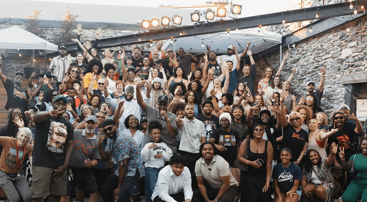 Large group of people gathered outdoors, some raising their hands, in front of a stone wall and stage lights.