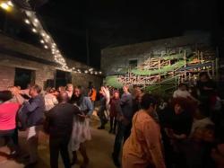 People dancing and socializing indoors with string lights and a large wooden structure in the background.