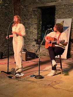A woman singing into a microphone and a man playing guitar on stage with brick wall background.