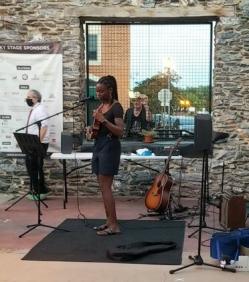 A young girl stands on a stage with a microphone, a guitar, and music equipment, performing in front of a stone wall and window.