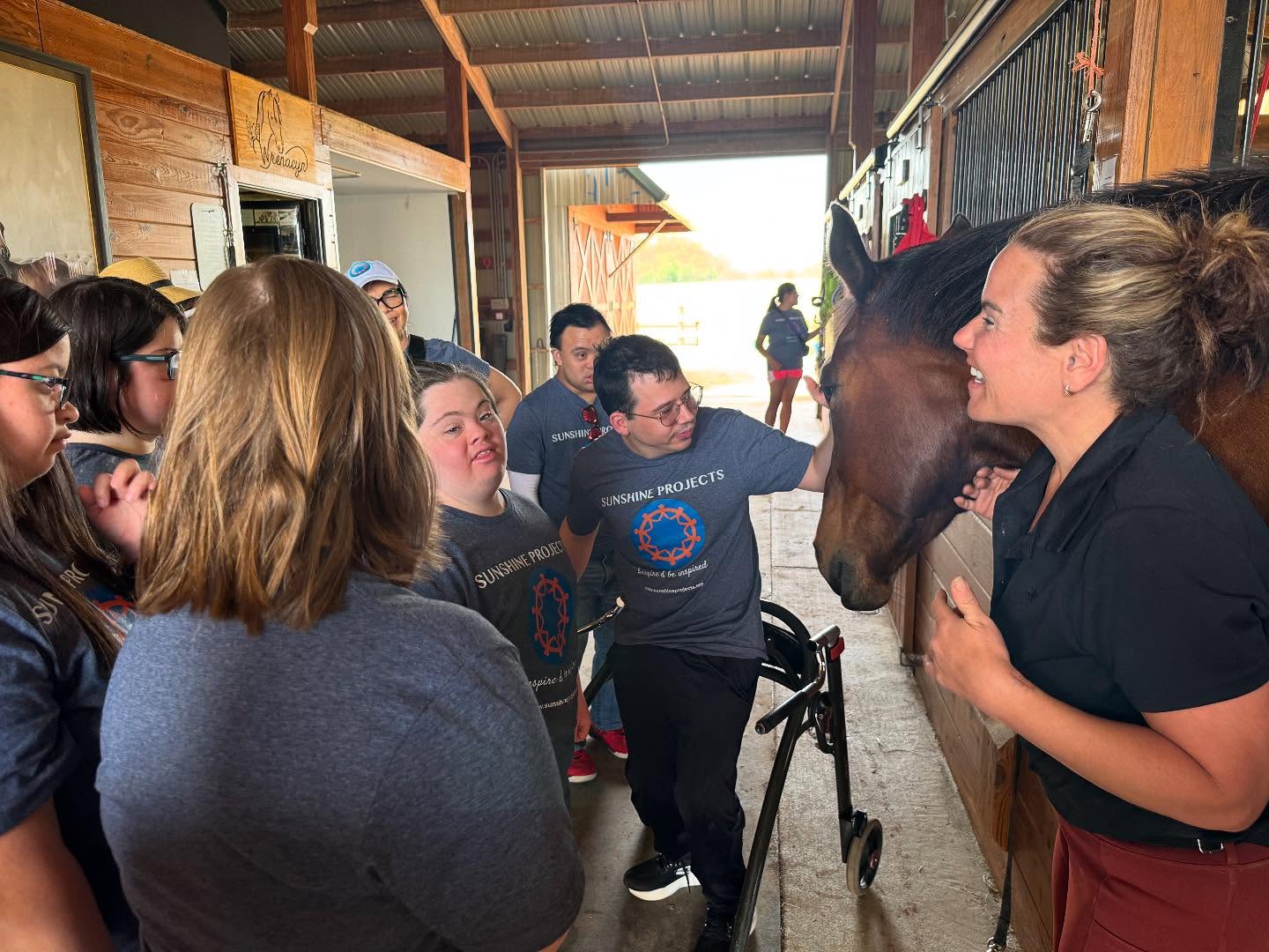 Group of people gathered around a woman and a horse in a stable, engaging in conversation.