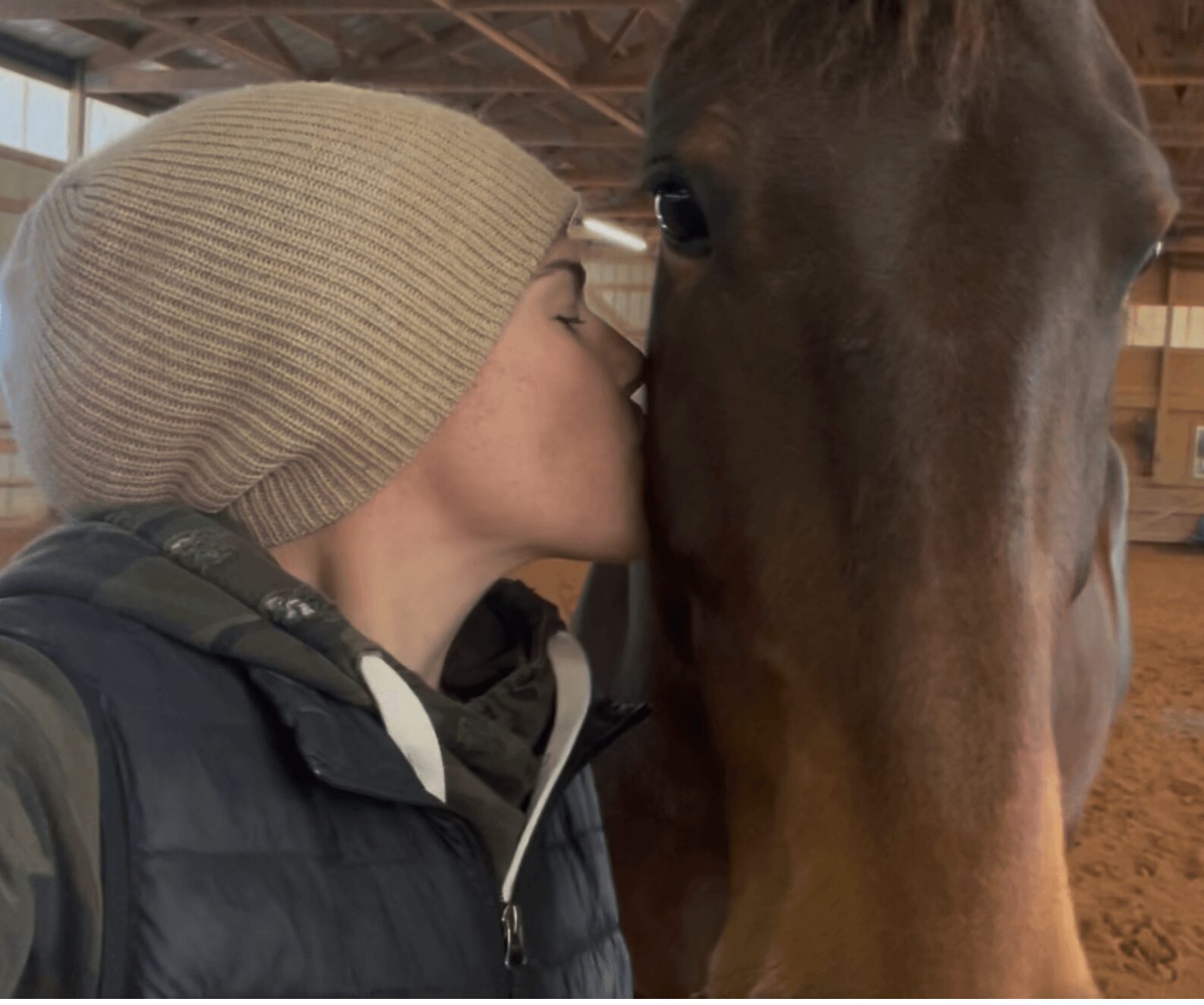 Person wearing a beige knit hat kisses a brown horse's face inside a stable.