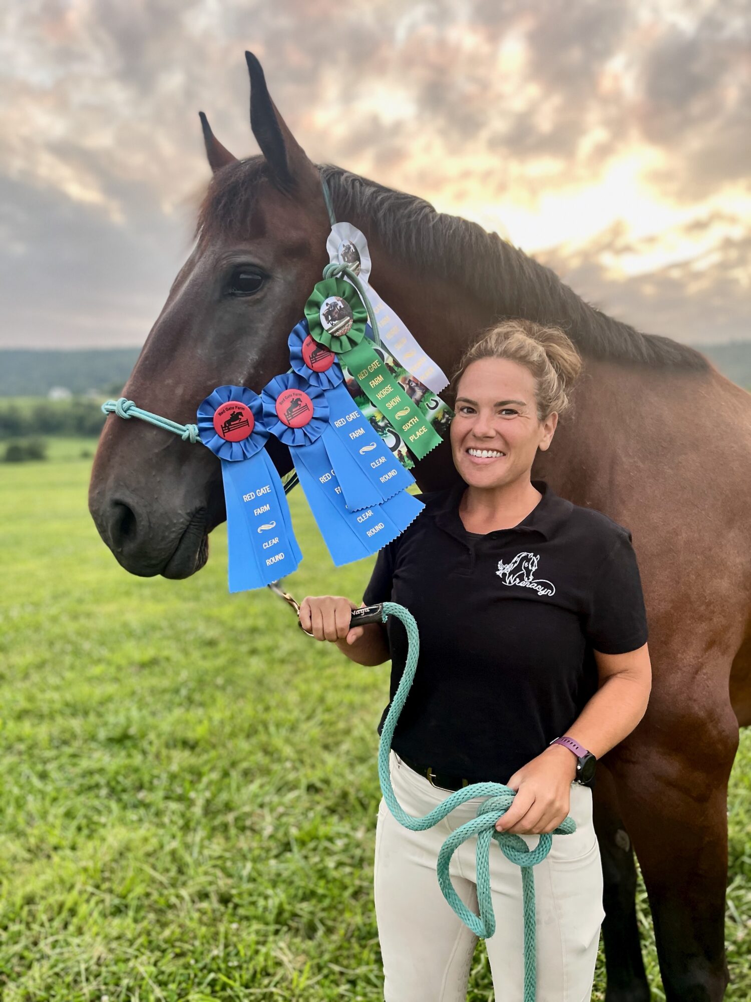 Woman smiling next to a horse with ribbons and medals in a grassy field during sunset