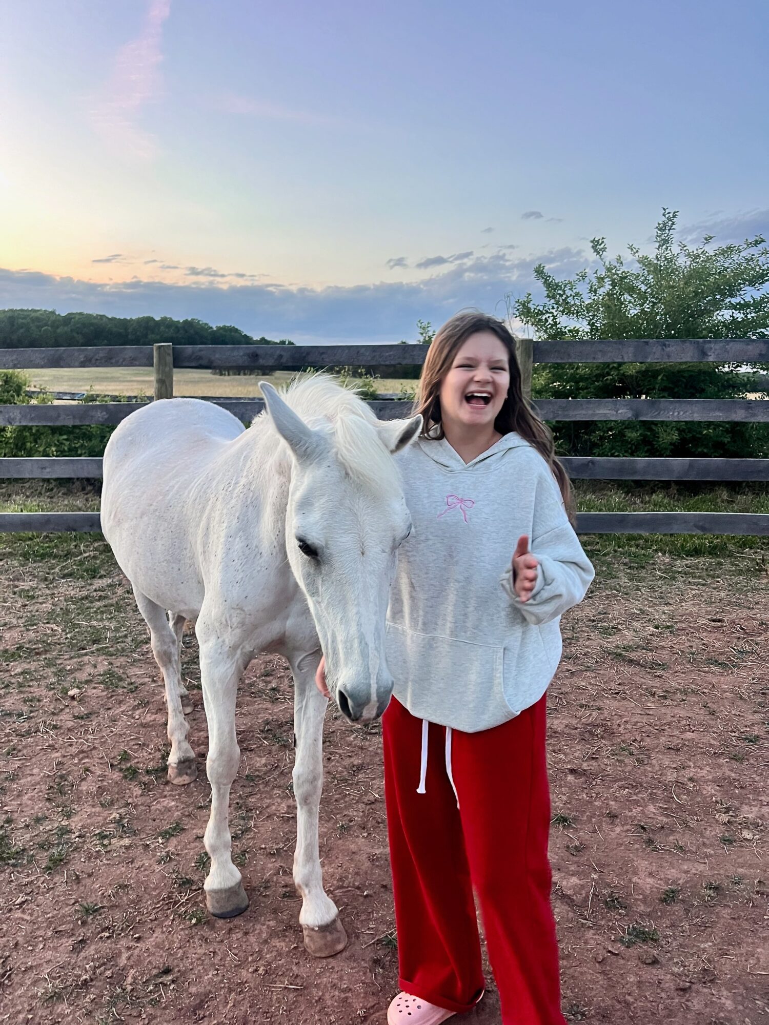 Girl smiling and giving a thumbs-up next to a white horse outdoors, with a fence and trees in the background.