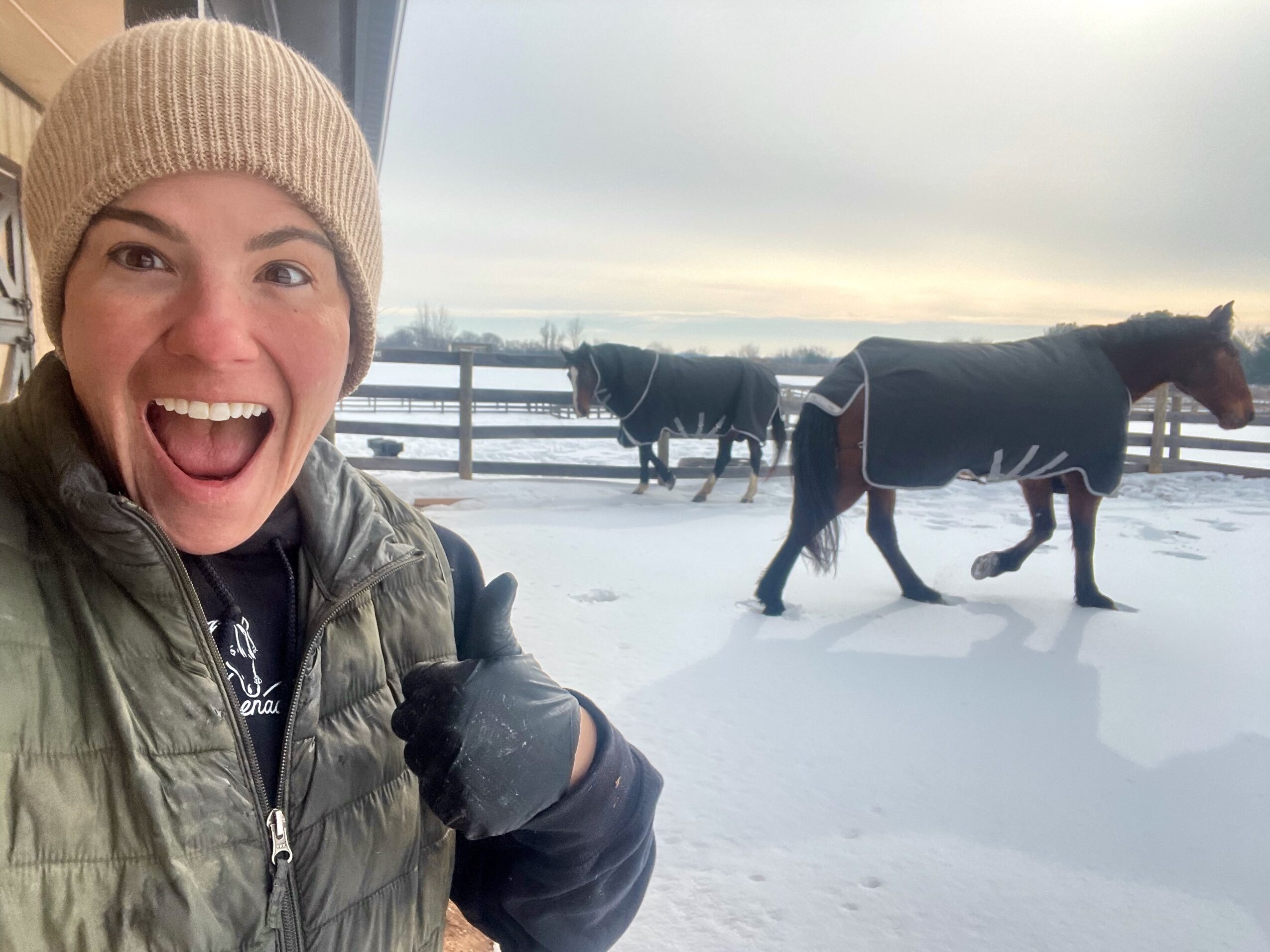 Woman smiling outdoors in winter, snow-covered ground, horses in the background, wearing a beanie and jacket.