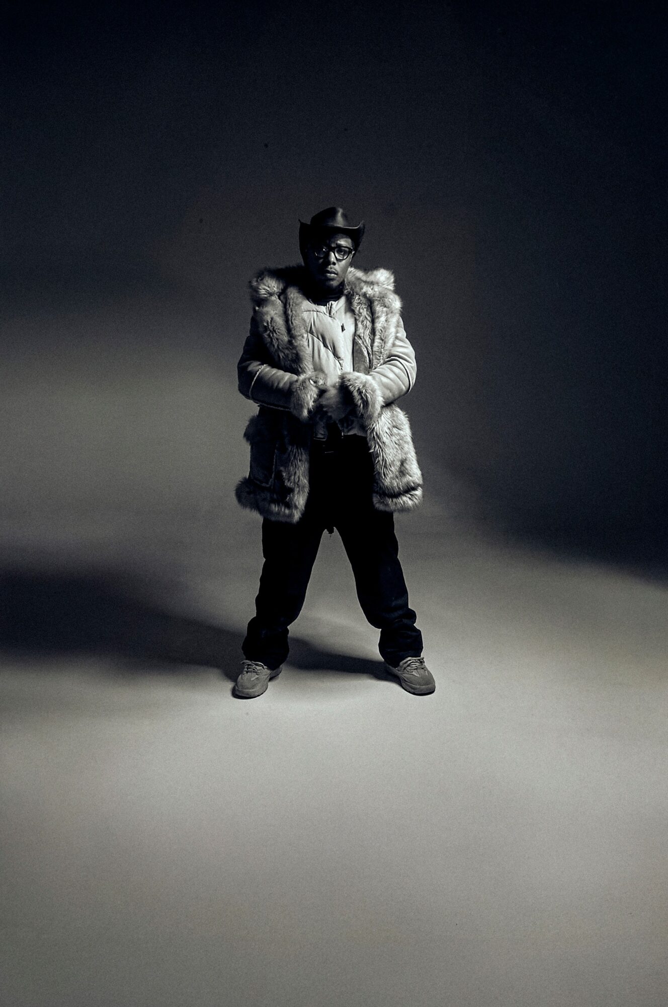Person standing in a spotlight, wearing a fur coat, hat, and sunglasses, with a dark background.