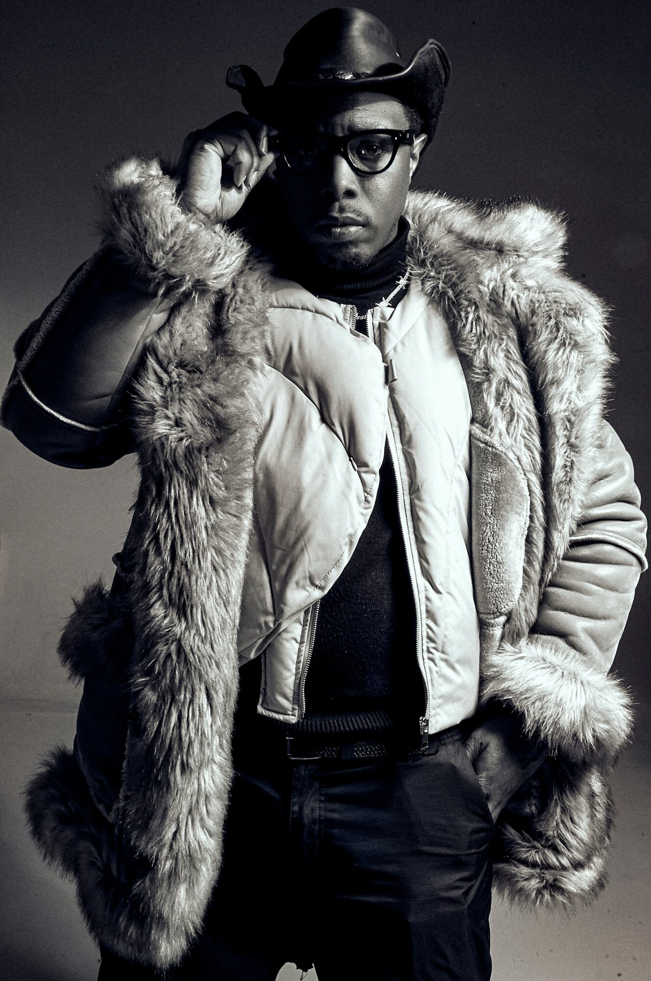 Person wearing glasses, a fur-lined coat, and a hat, adjusting glasses with one hand, in black and white.