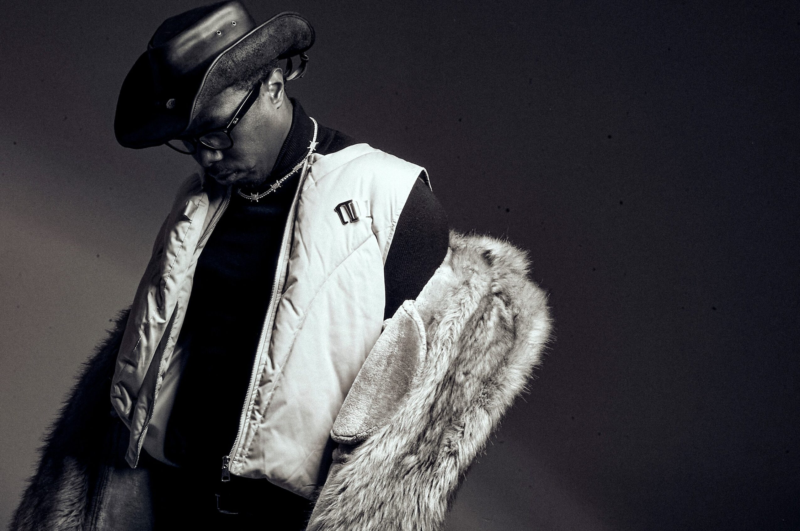 Person wearing a hat, glasses, a vest, and a fur coat draped over shoulders, looking down, in black and white.