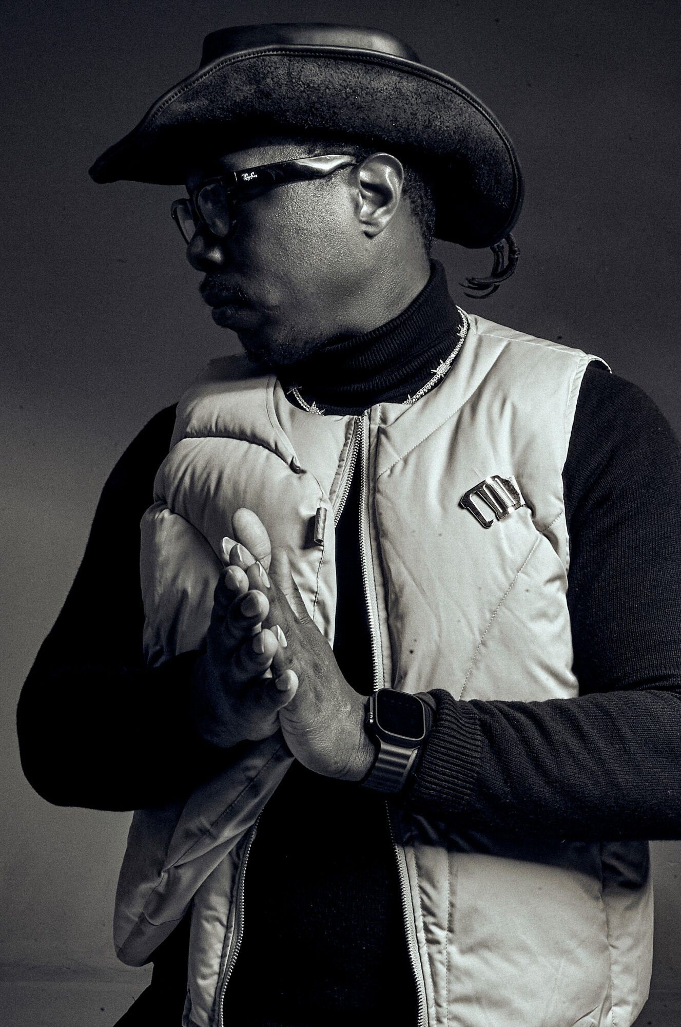 Person wearing a cowboy hat, glasses, and a puffy vest, with hands clasped together, in black and white.