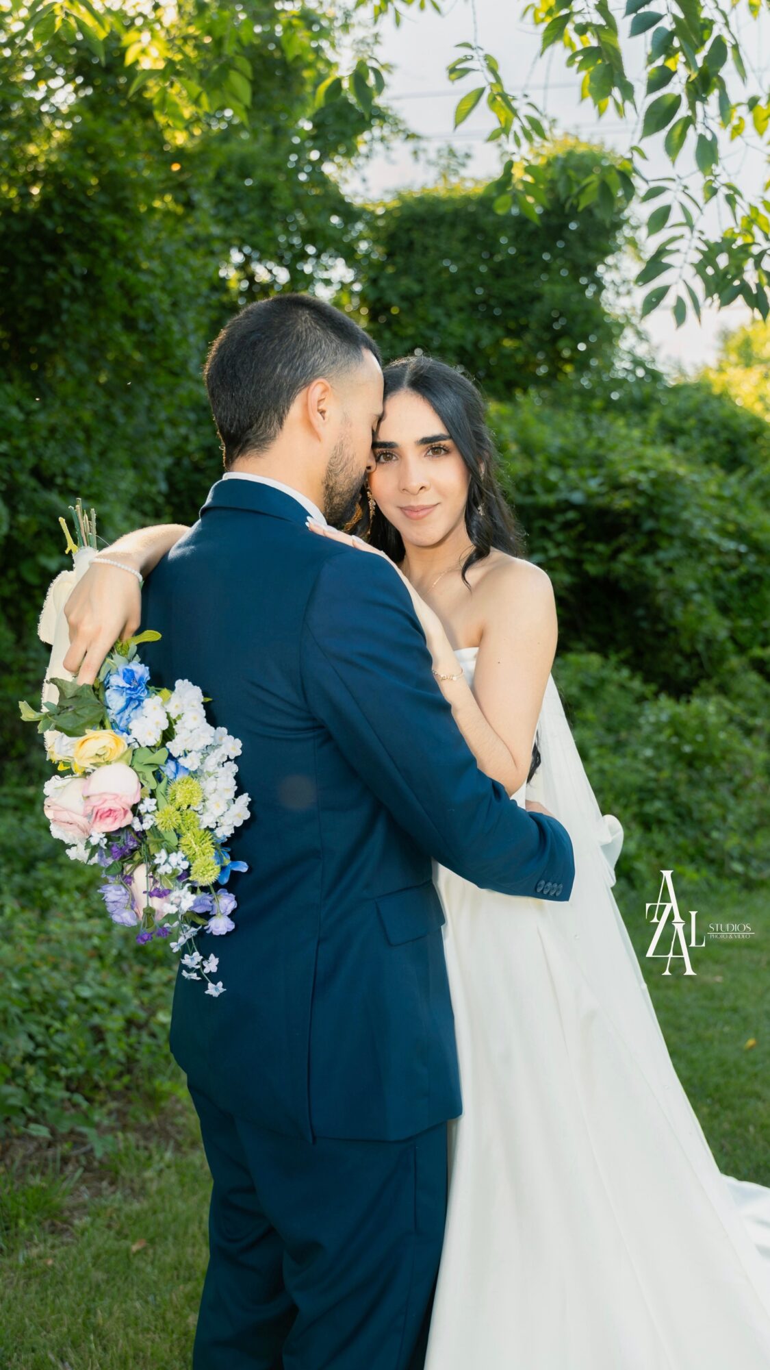 A bride and groom embrace outdoors with greenery in the background, the bride holding a bouquet of flowers.