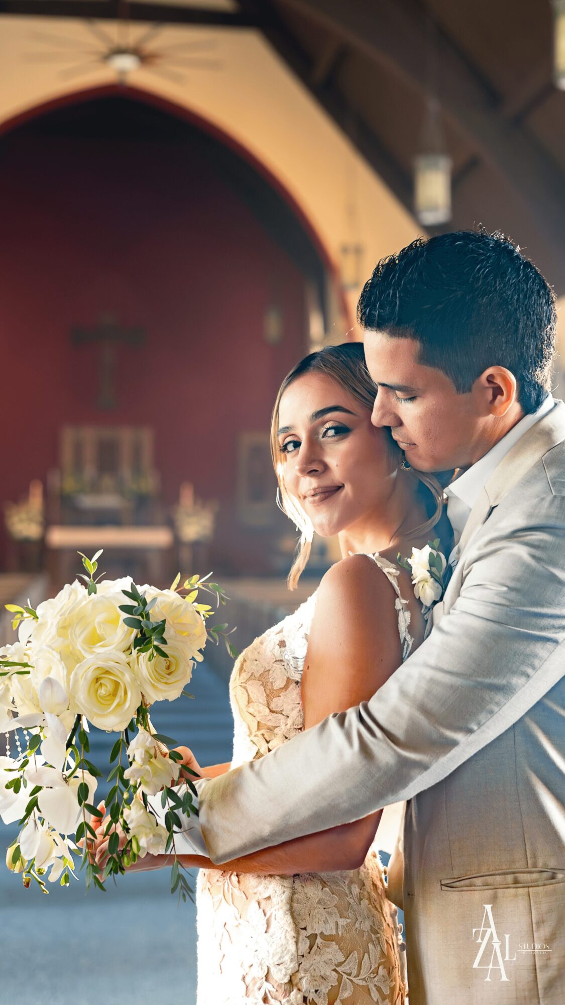 A couple in wedding attire embraces, with the bride holding a bouquet of white flowers, inside a church with a red arch.