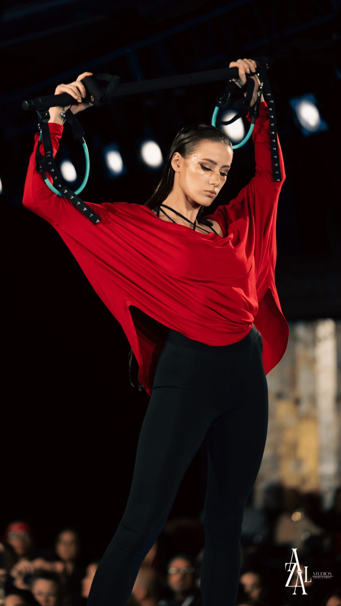 Woman in red top and black pants holding gymnastic rings overhead, focused expression, dark background.