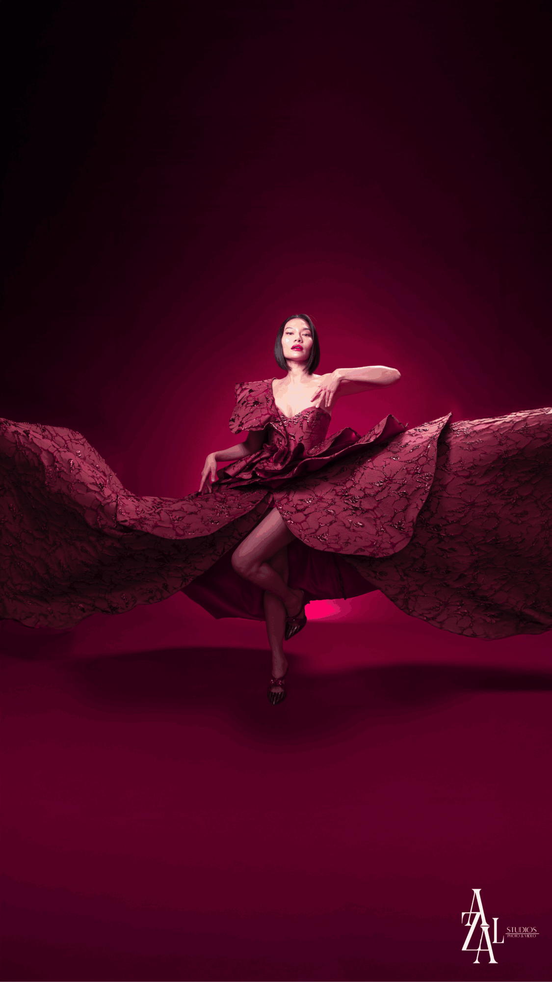 A woman in a dress performs a dance move on a stage with a dark pink background.