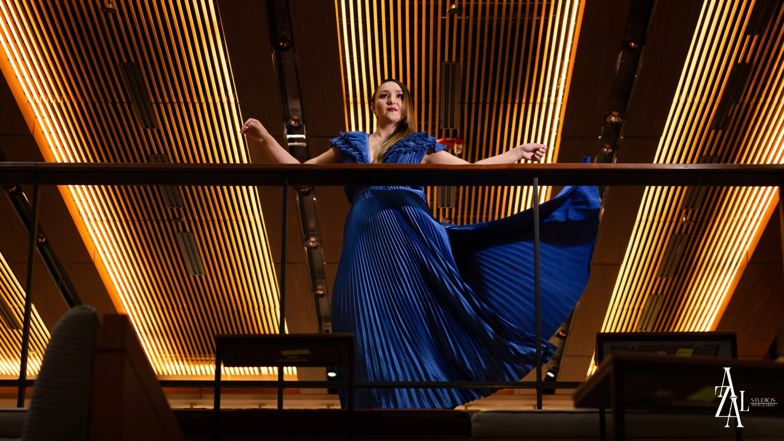 Woman in a blue dress standing on a balcony with golden vertical light fixtures behind her.
