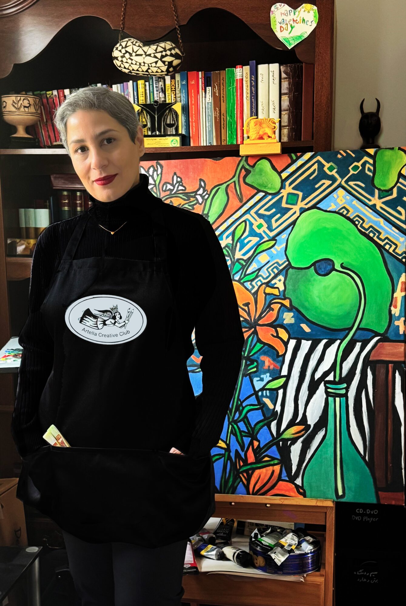 Woman with dark hair and red lipstick standing indoors in front of colorful artwork and bookshelves.