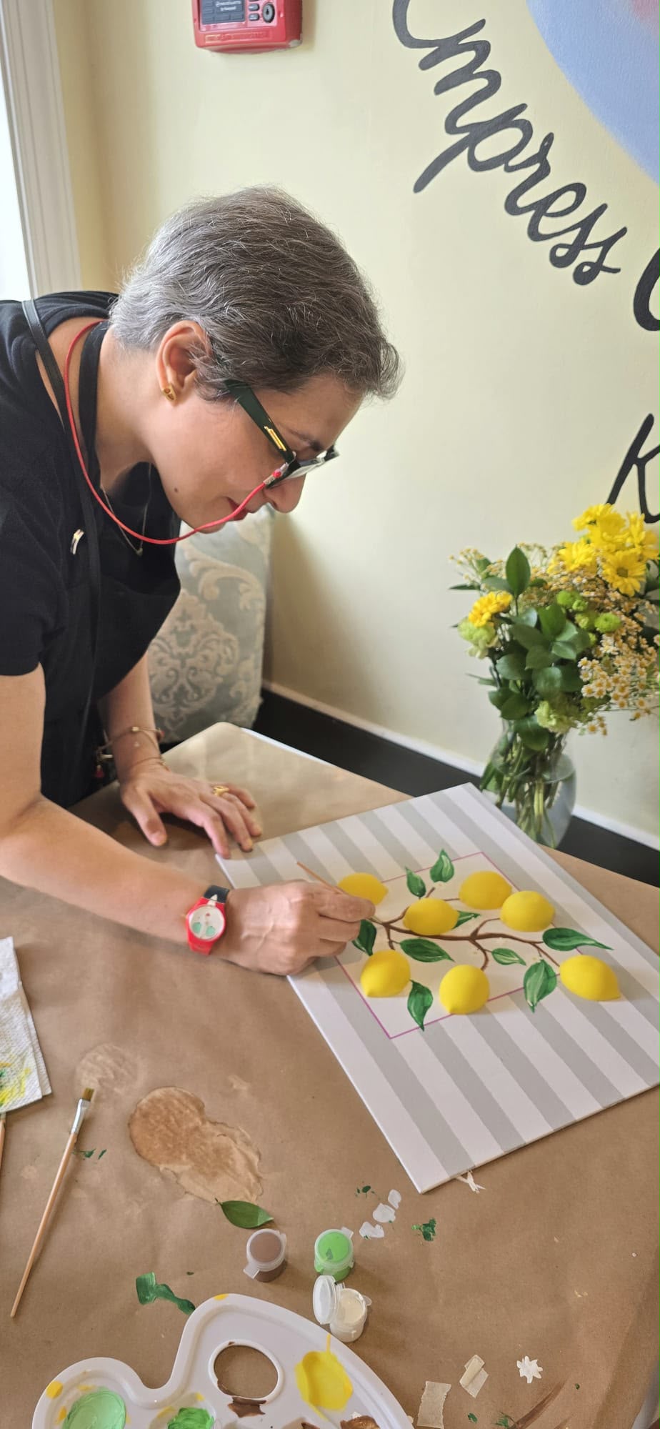 Elderly woman decorating cookies with yellow and green icing on a striped surface, with a vase of yellow flowers nearby.
