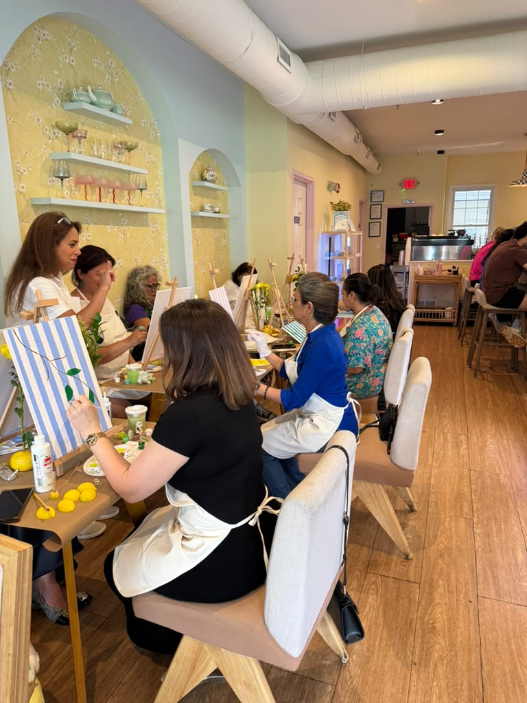 People sitting at a table in a decorated room, some painting, with pastel-colored walls and wooden floors.