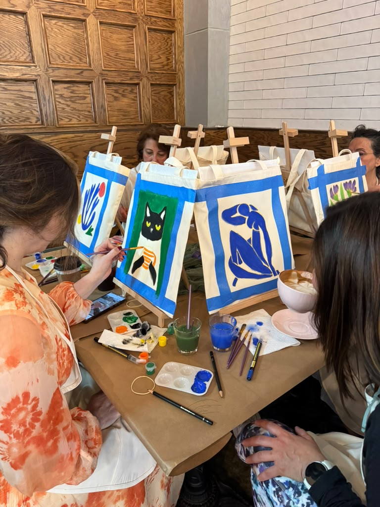 People painting animal-themed pictures on fabric panels, with art supplies on a table, in a room with wooden and brick walls.