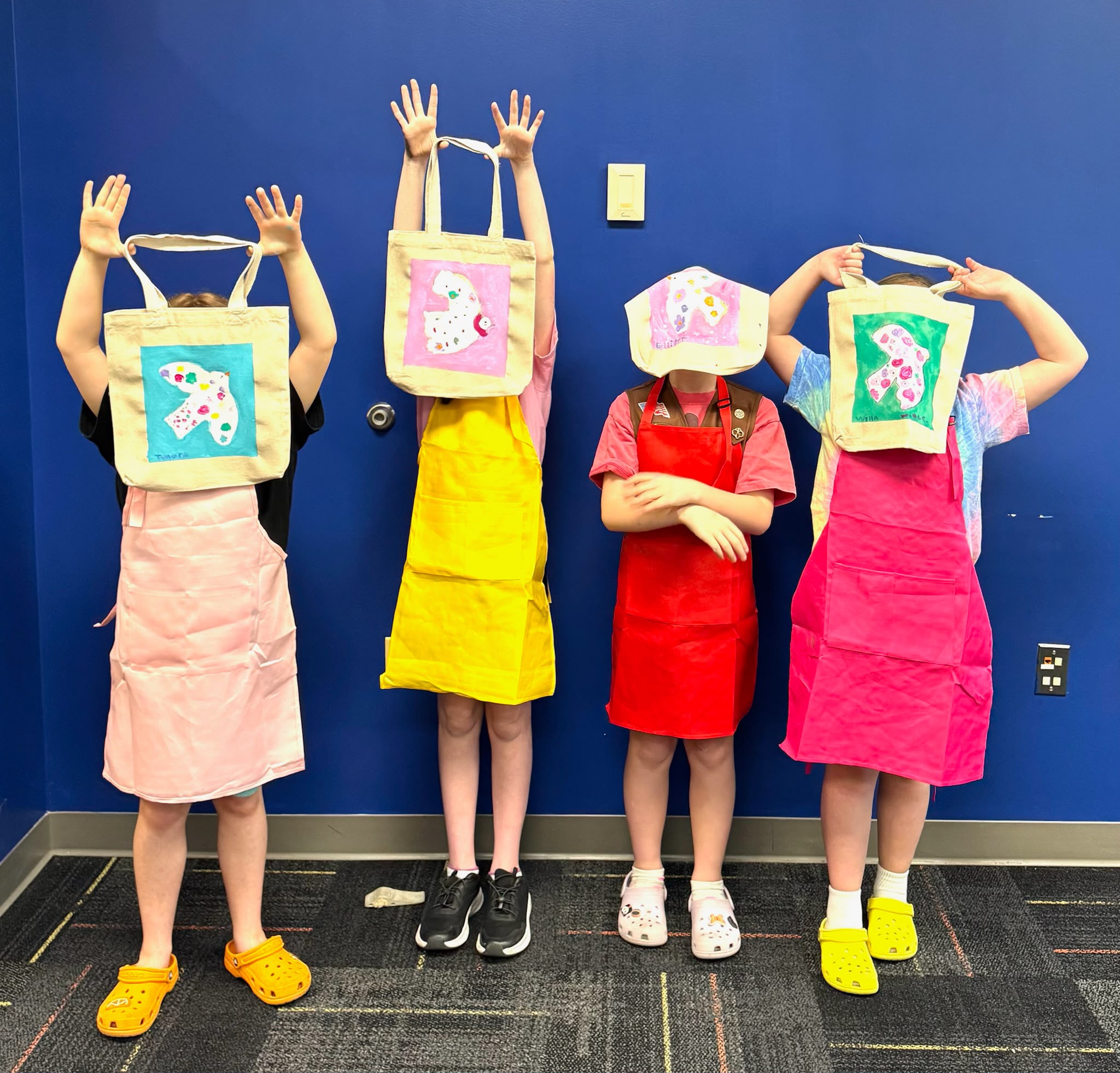 Four children stand against a blue wall, wearing colorful aprons and unicorn-themed bags over their heads, posing playfully.