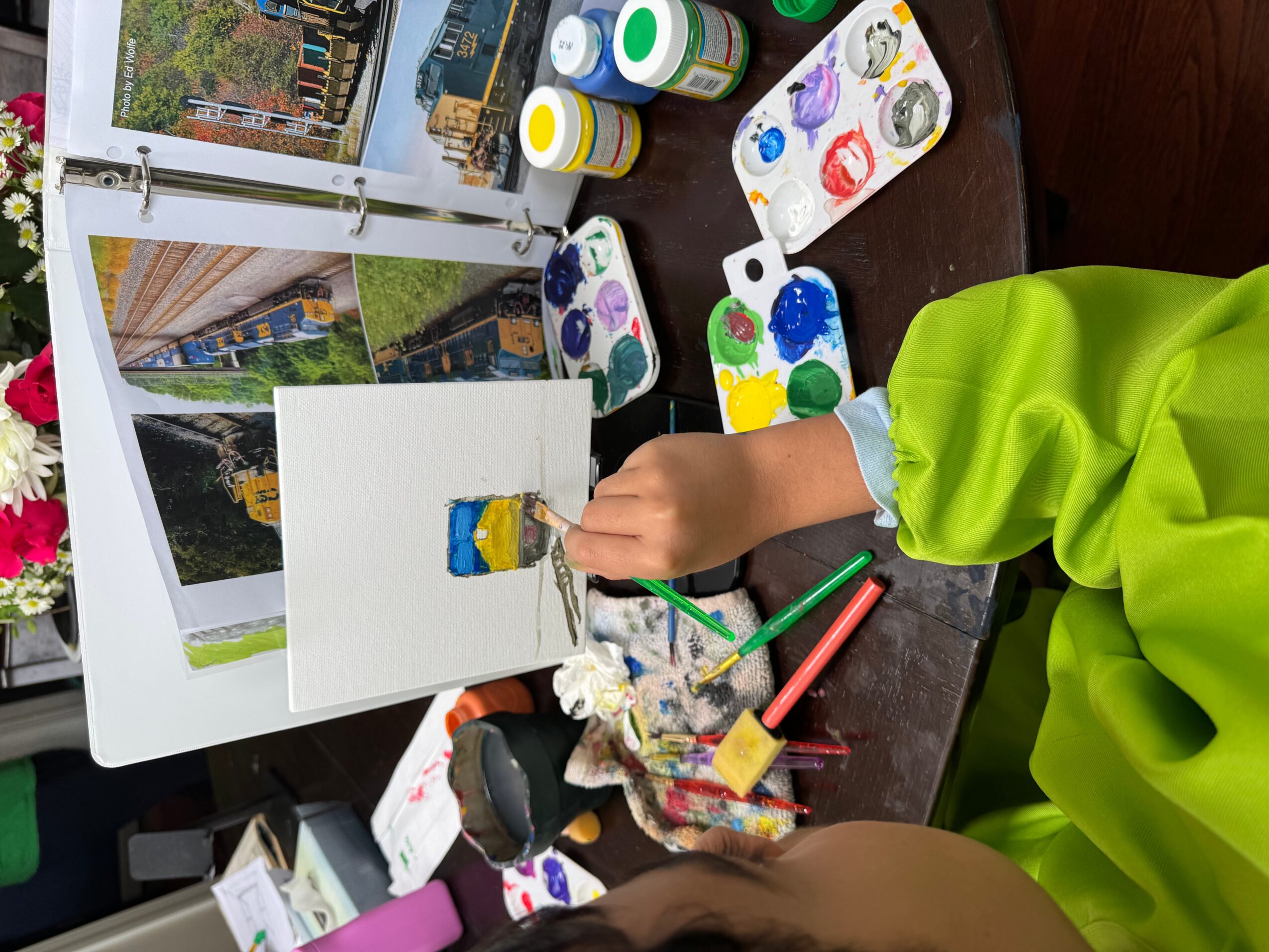 Child in green shirt painting a colorful ice cream cone on paper, surrounded by paint supplies and pictures, on a dark table.