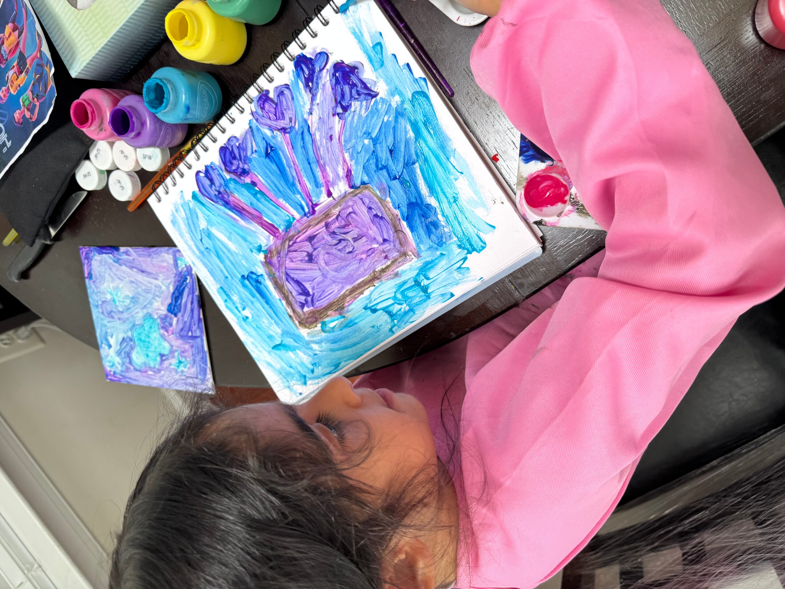Child in pink smock painting with blue and purple colors on paper, surrounded by paint bottles and art supplies.