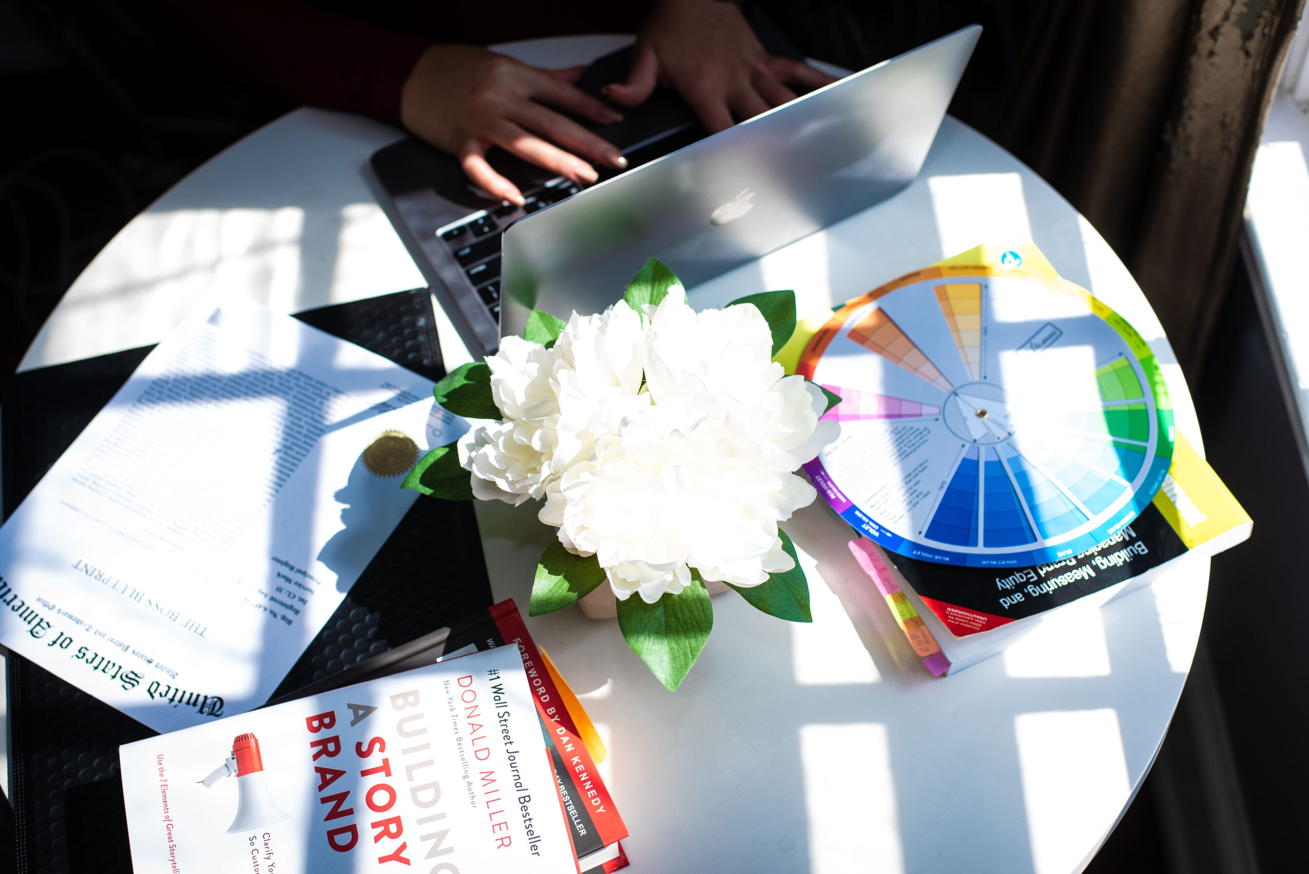 Hands typing on a laptop with documents, a flower arrangement, and a colorful chart on a white table.