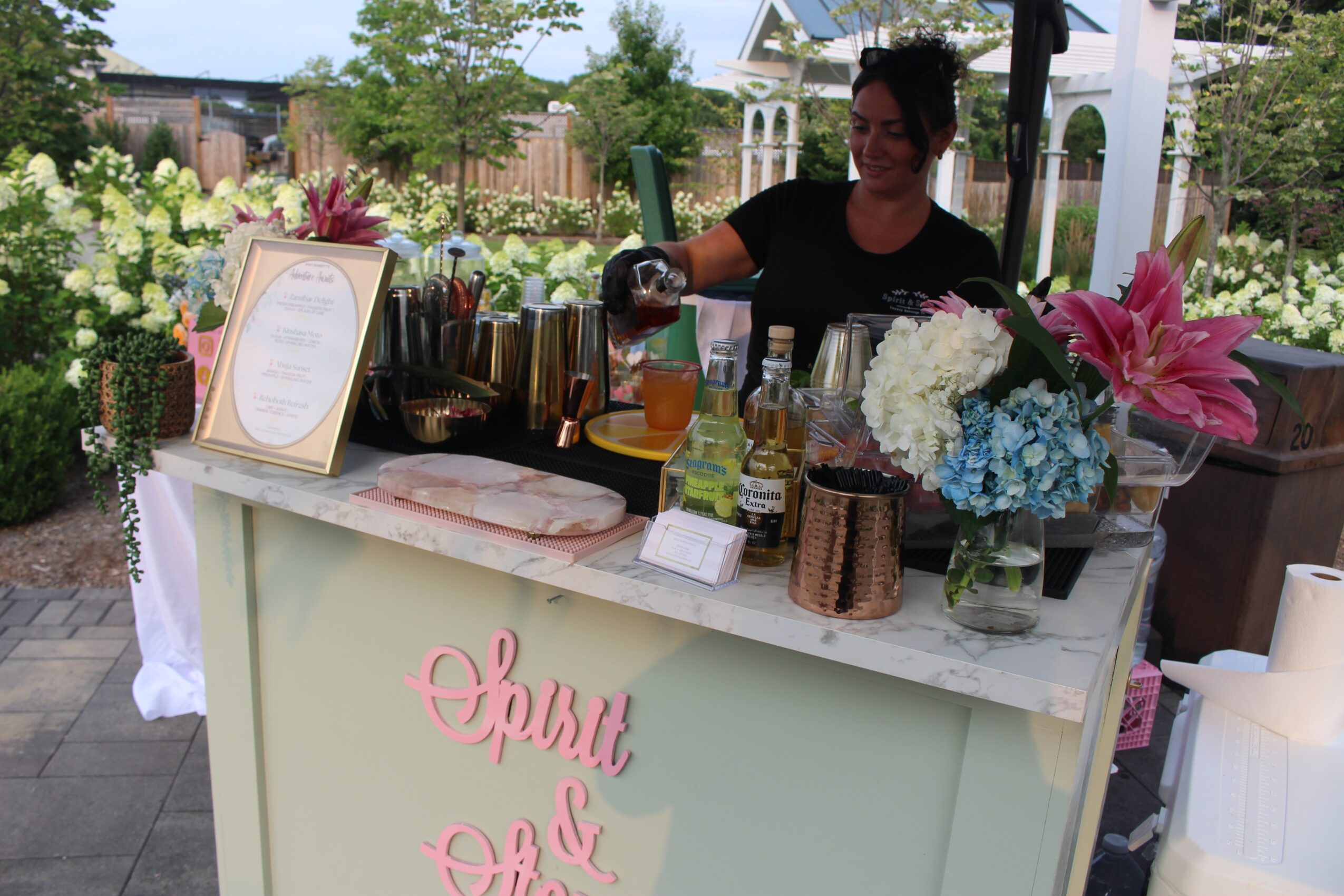 Person preparing drinks at an outdoor bar with flowers and bottles, garden background, sunny day.