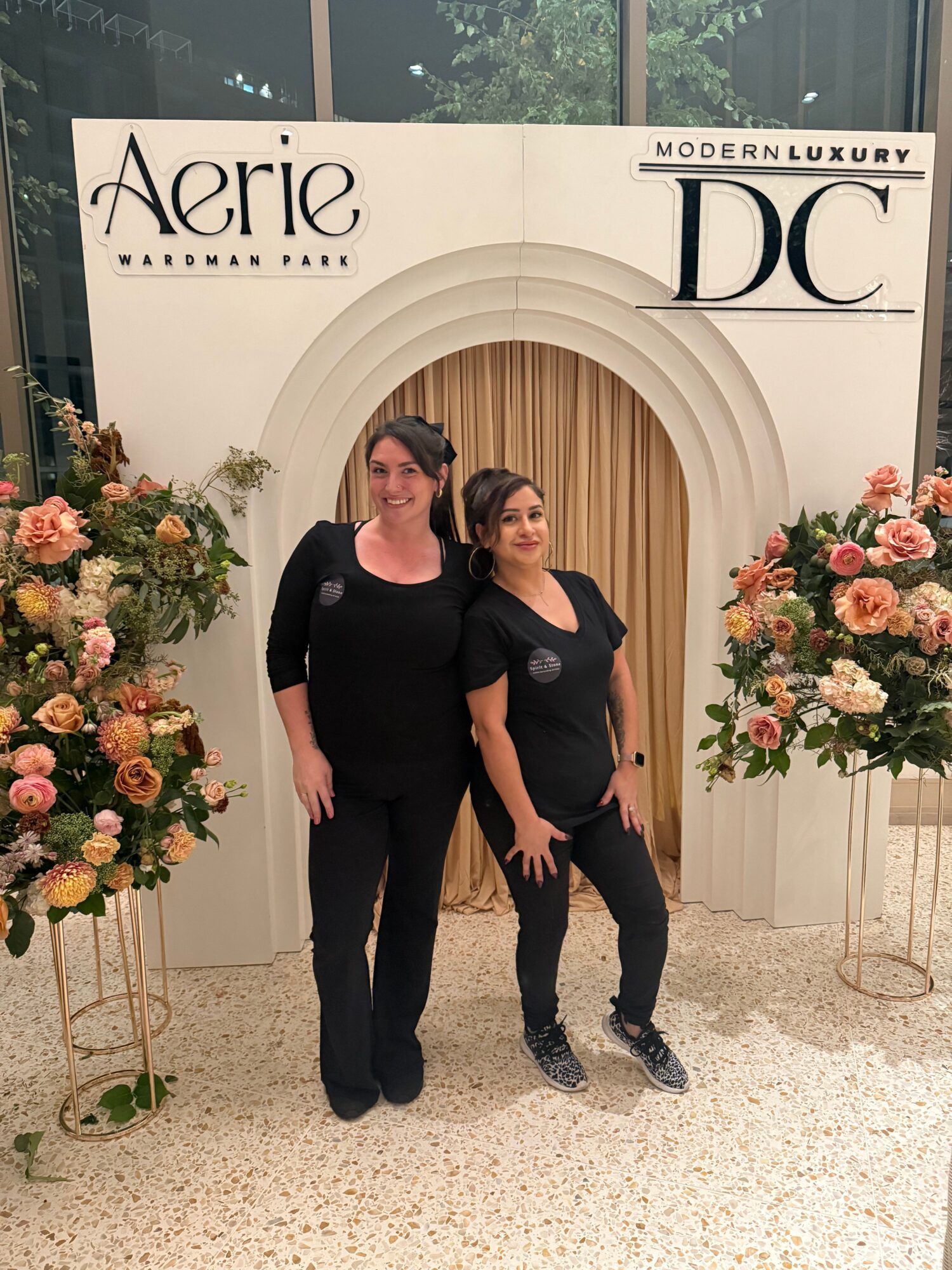 Two women standing in front of a white backdrop with floral arrangements, one in black and the other in dark clothing.