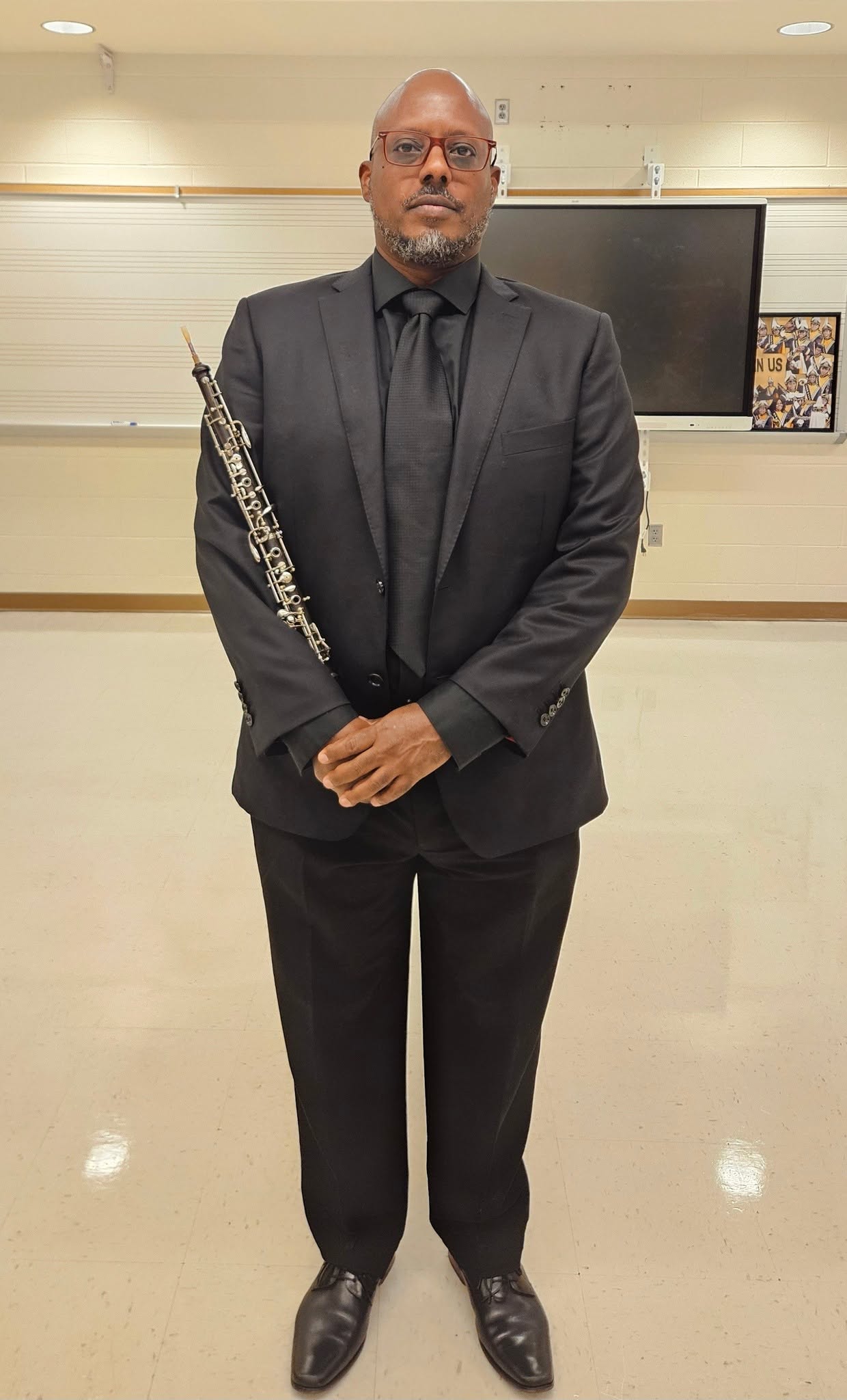 Man in black suit holding a flute, standing in a room with a large screen behind him.