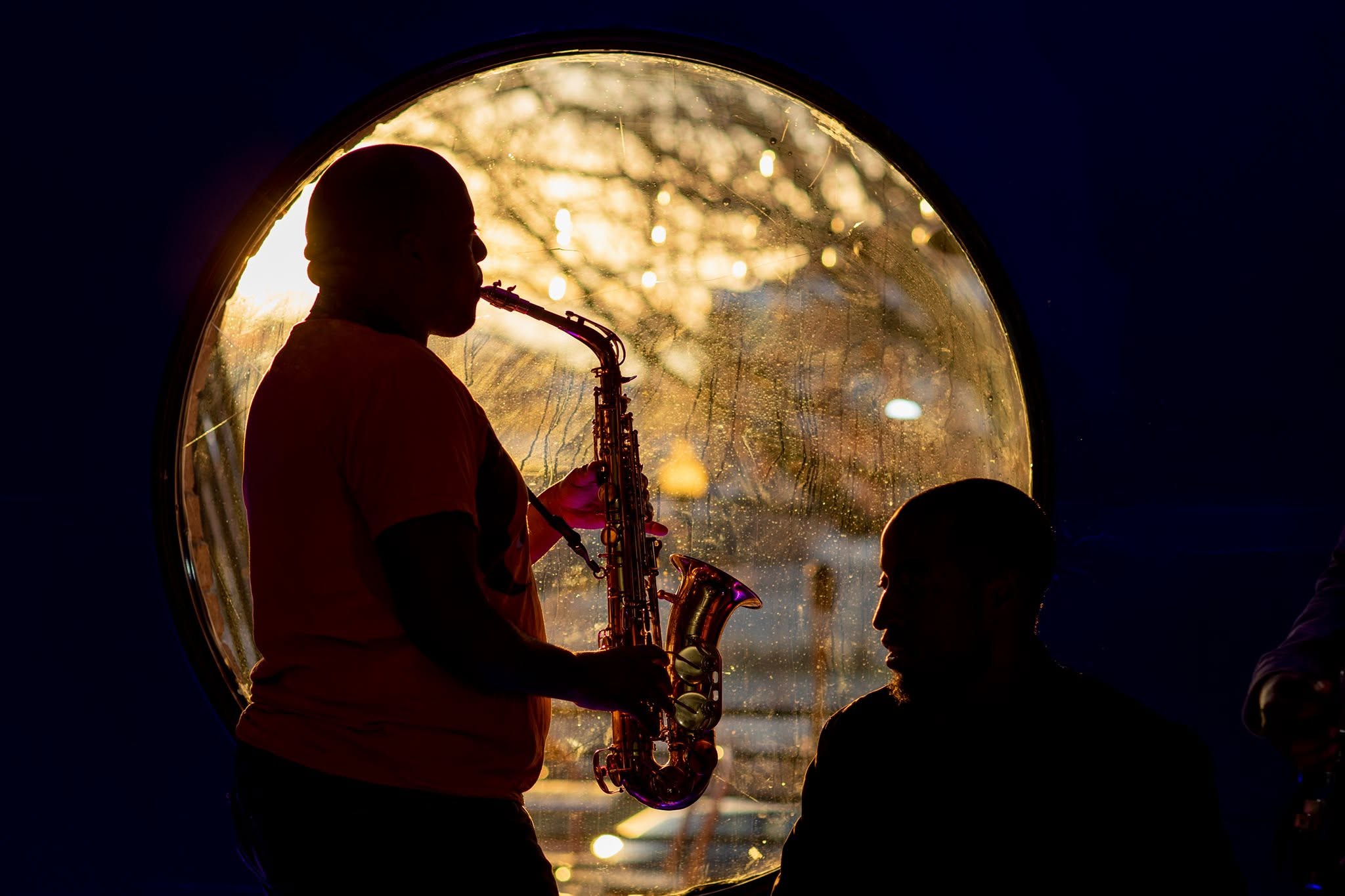 Silhouette of a person playing a saxophone near a circular window with a sunset background.