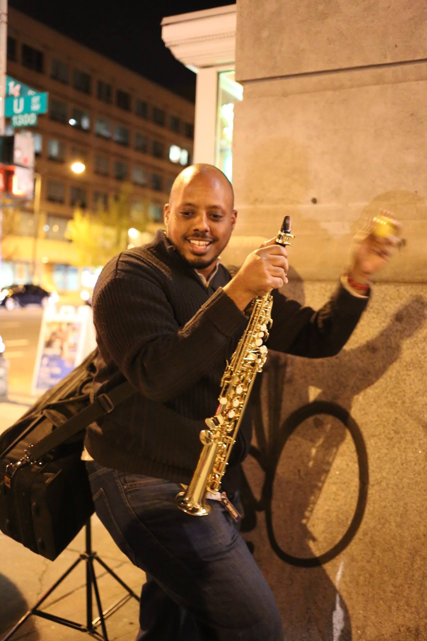Smiling man holding a saxophone outdoors at night, standing next to a wall with a bicycle shadow, city lights in background.