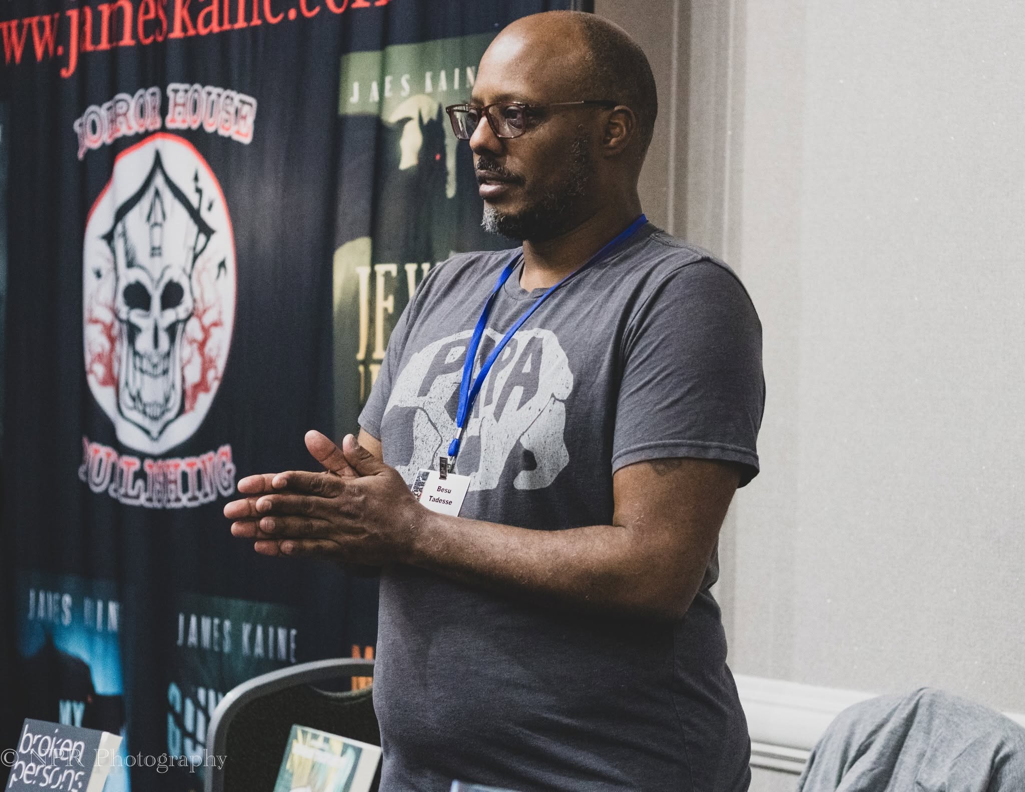 Man with glasses and a lanyard stands indoors, wearing a dark T-shirt, with banners in the background.