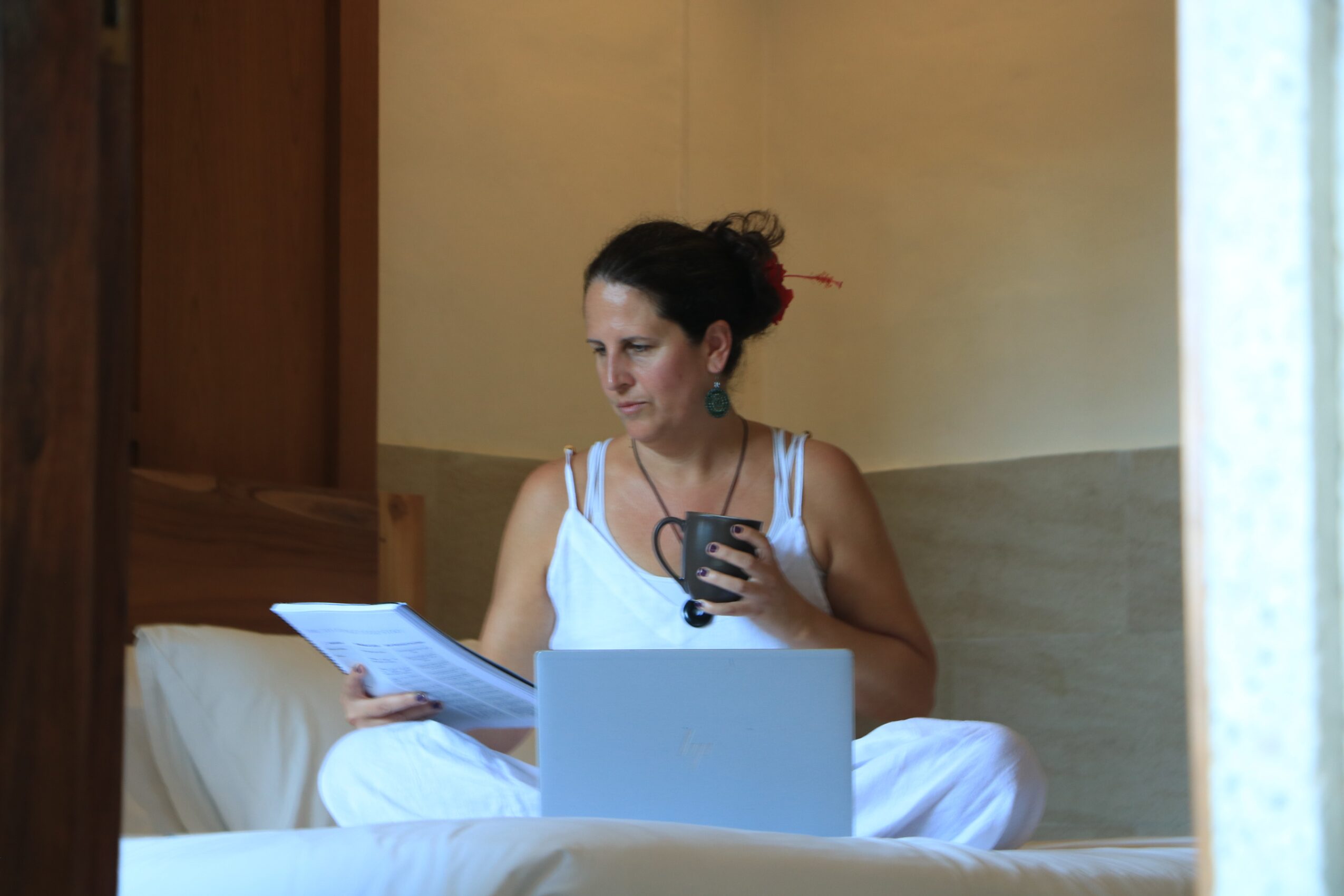 Woman sitting on bed, reading a document and holding a phone, with a laptop in front of her.