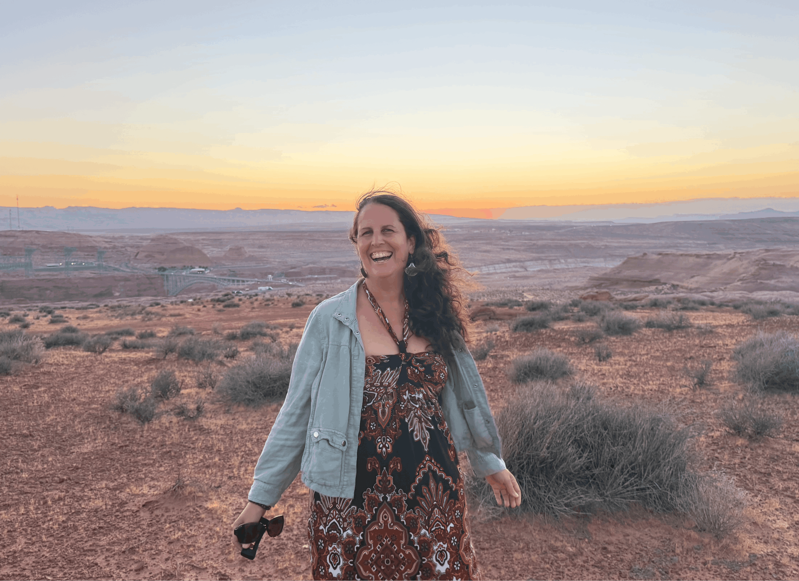 Woman standing outdoors at sunset, smiling, with desert landscape and distant horizon behind her.